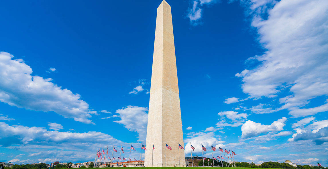 Washington 桌布 壁紙 — 高聳的華盛頓紀念碑在點綴著蓬鬆白雲的鮮豔藍天下巍然矗立，周圍環繞著飄揚的美國國旗。