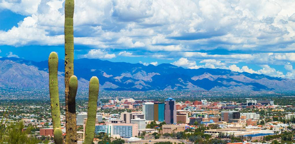Arizona 桌布 壁紙 — 高聳的薩瓜羅仙人掌框住一個廣闊的沙漠城市，擁有現代建築和遠處的山脈，在戲劇性的多雲天空下。