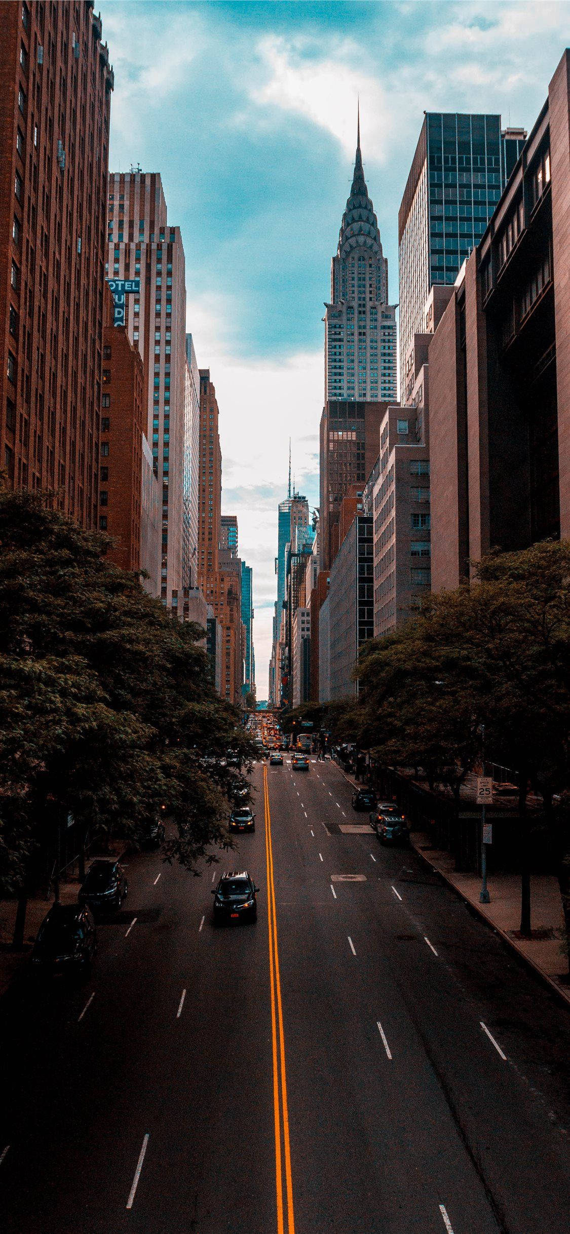 New York 桌布 壁紙 — 高聳的都市峽谷，兩旁摩天大樓林立，克萊斯勒大廈在樹蔭大道上方穿透部分多雲的天空。