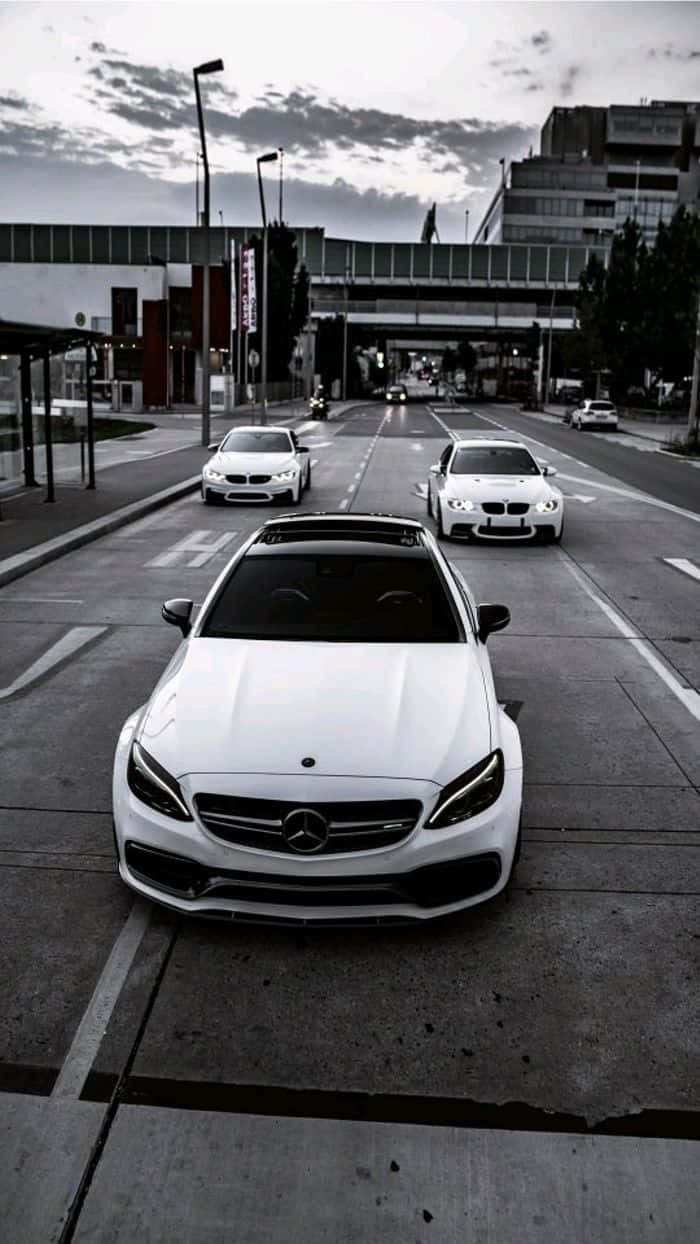 Mercedes 桌布 壁紙 — 高角度拍攝一輛白色賓士轎車帶領另外兩輛豪華汽車在陰天下行駛於城市街道。