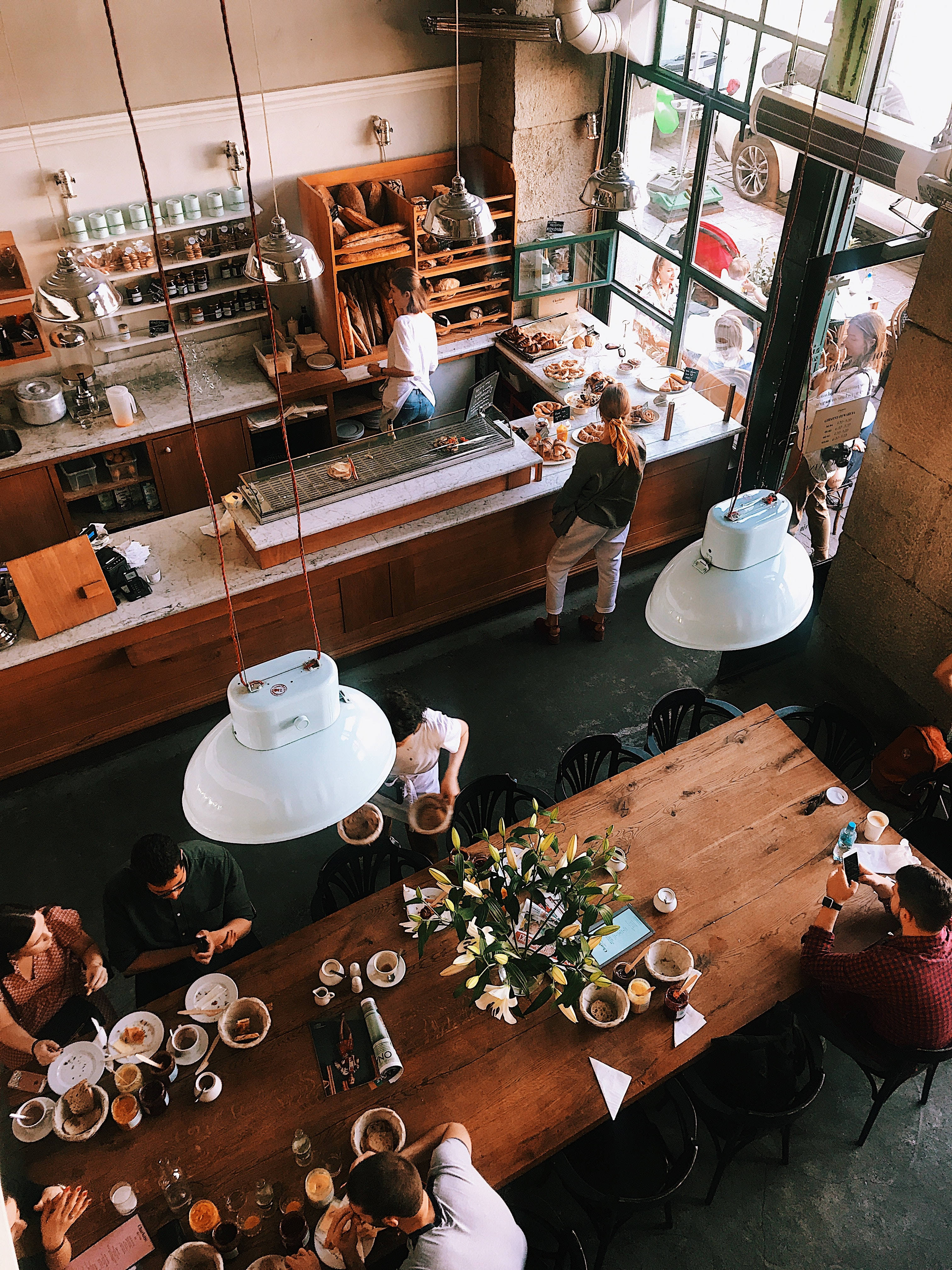 Coffee Shop 桌布 壁紙 — 高角度拍攝一間繁忙的咖啡館，顧客在大型白色吊燈下的長木桌上用餐。