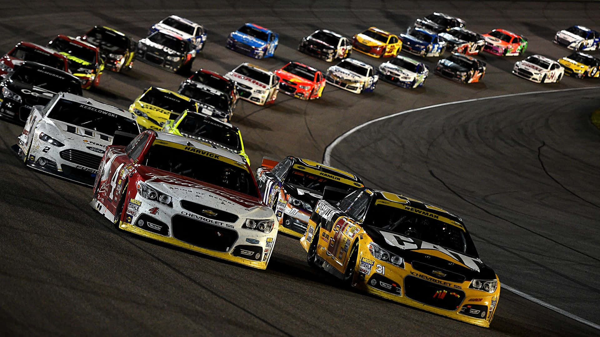 nascar 桌布 壁紙 — 高速夜間NASCAR賽事，色彩鮮豔的量產車在人工燈光下緊密排列在彎曲賽道上。
