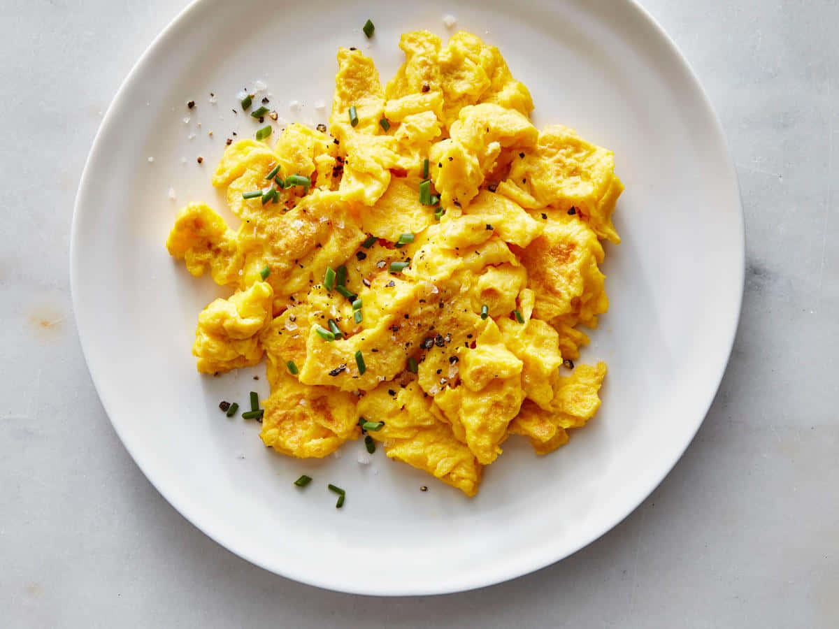 Scrambled Egg 桌布 壁紙 — 鬆軟金黃色炒蛋，飾以細香蔥和胡椒，放在白色盤子上，置於淺灰色表面上。