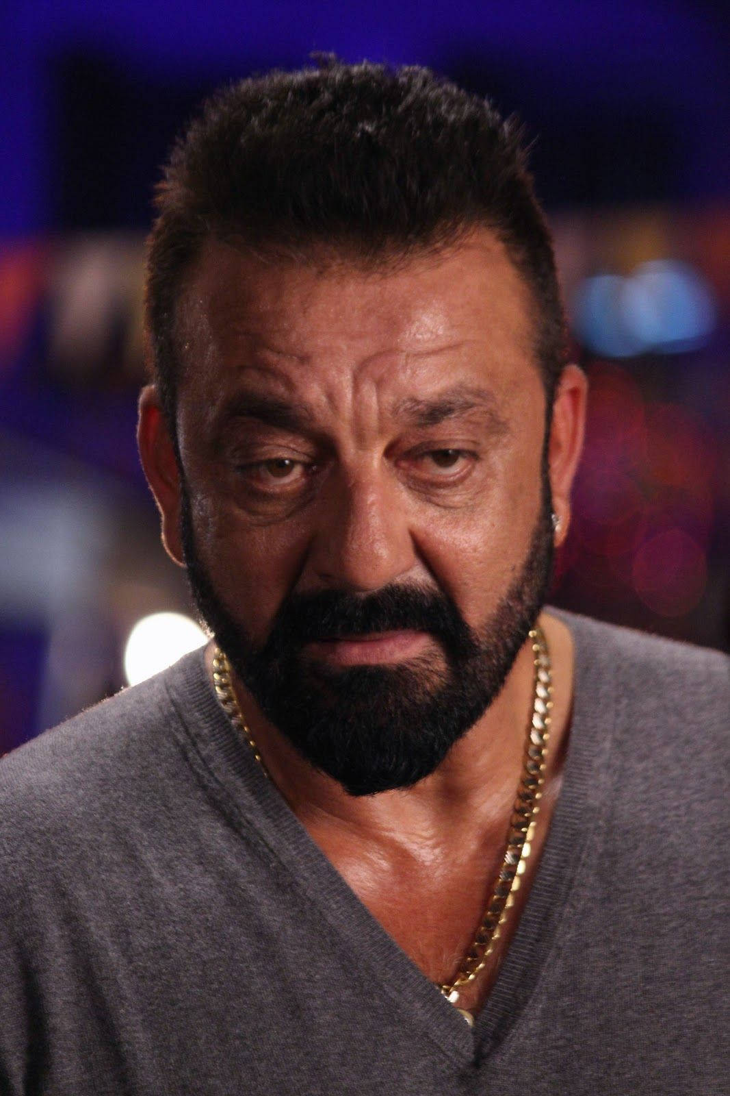 Sanjay 桌布 壁紙 — 鬍鬚男特寫肖像，眼神專注，身穿灰色V領上衣，戴金鏈，背景模糊多彩。