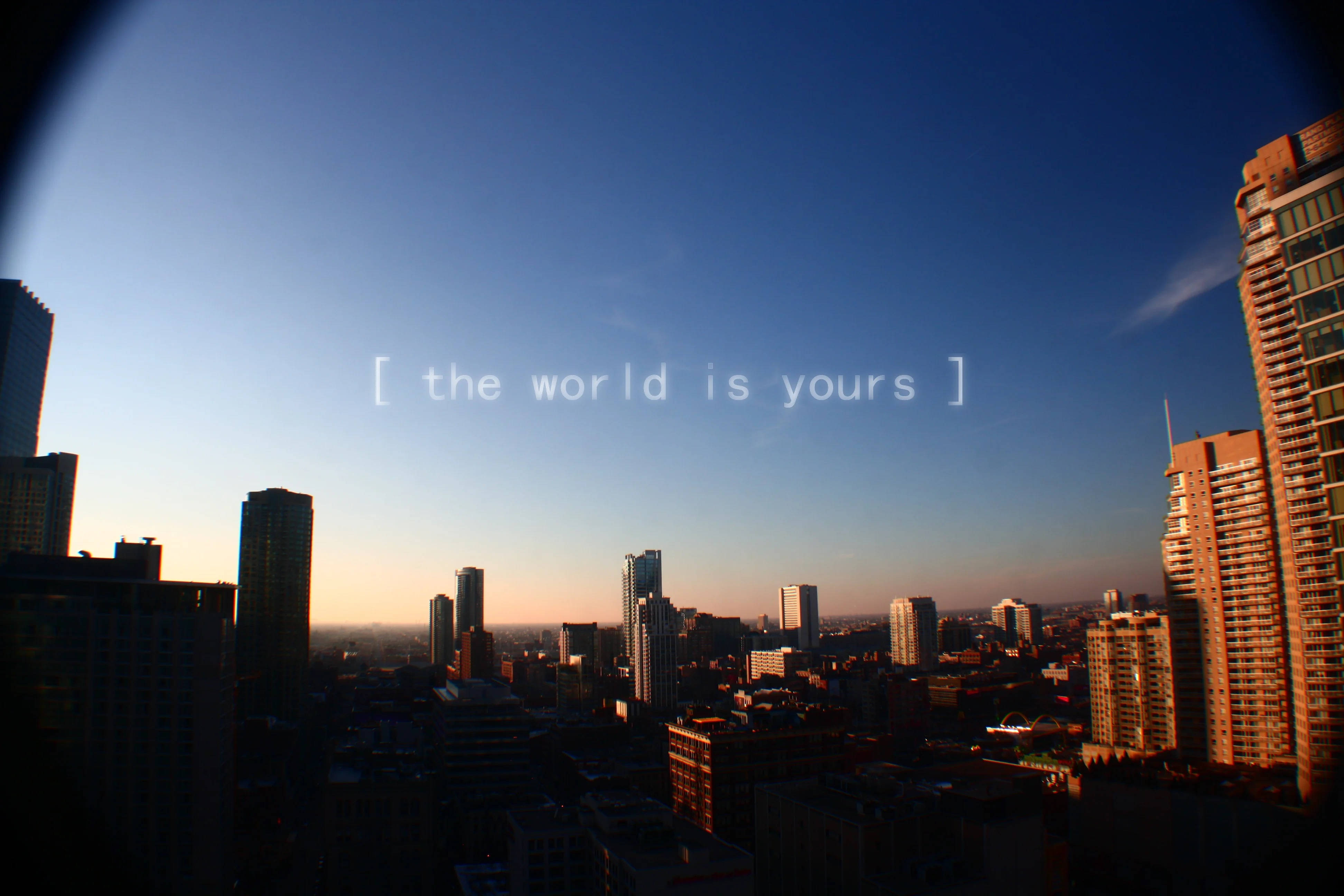 The World Is Yours 桌布 壁紙 — 魚眼鏡頭下的城市天際線日落景觀，摩天大樓上溫暖的金色光芒，漸變藍色天空上寫著「世界是你的」。