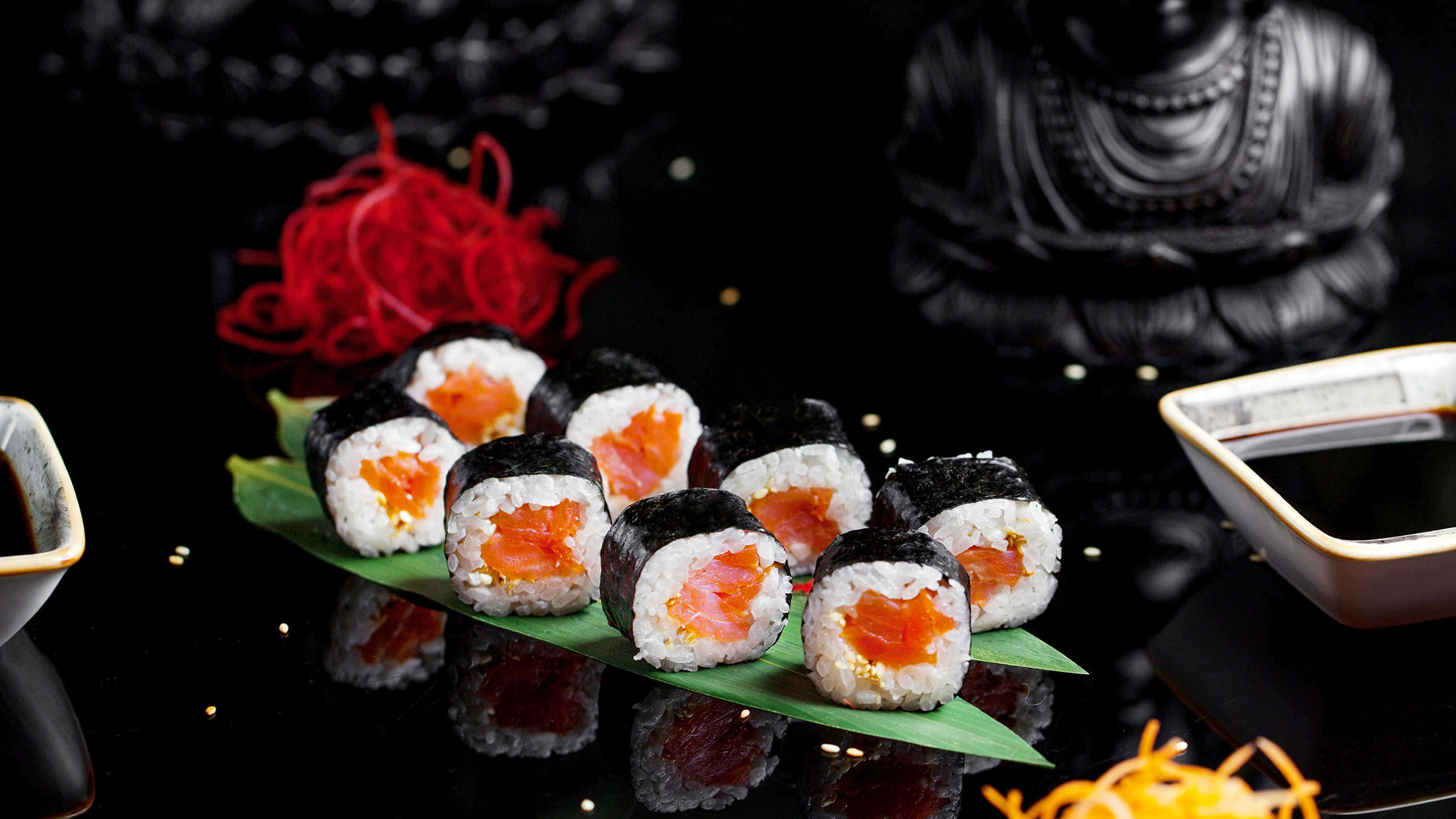 Sushi 桌布 壁紙 — 鮭魚壽司卷配飯放在綠葉上，置於黑色反光表面，配有紅色裝飾和醬油碗。