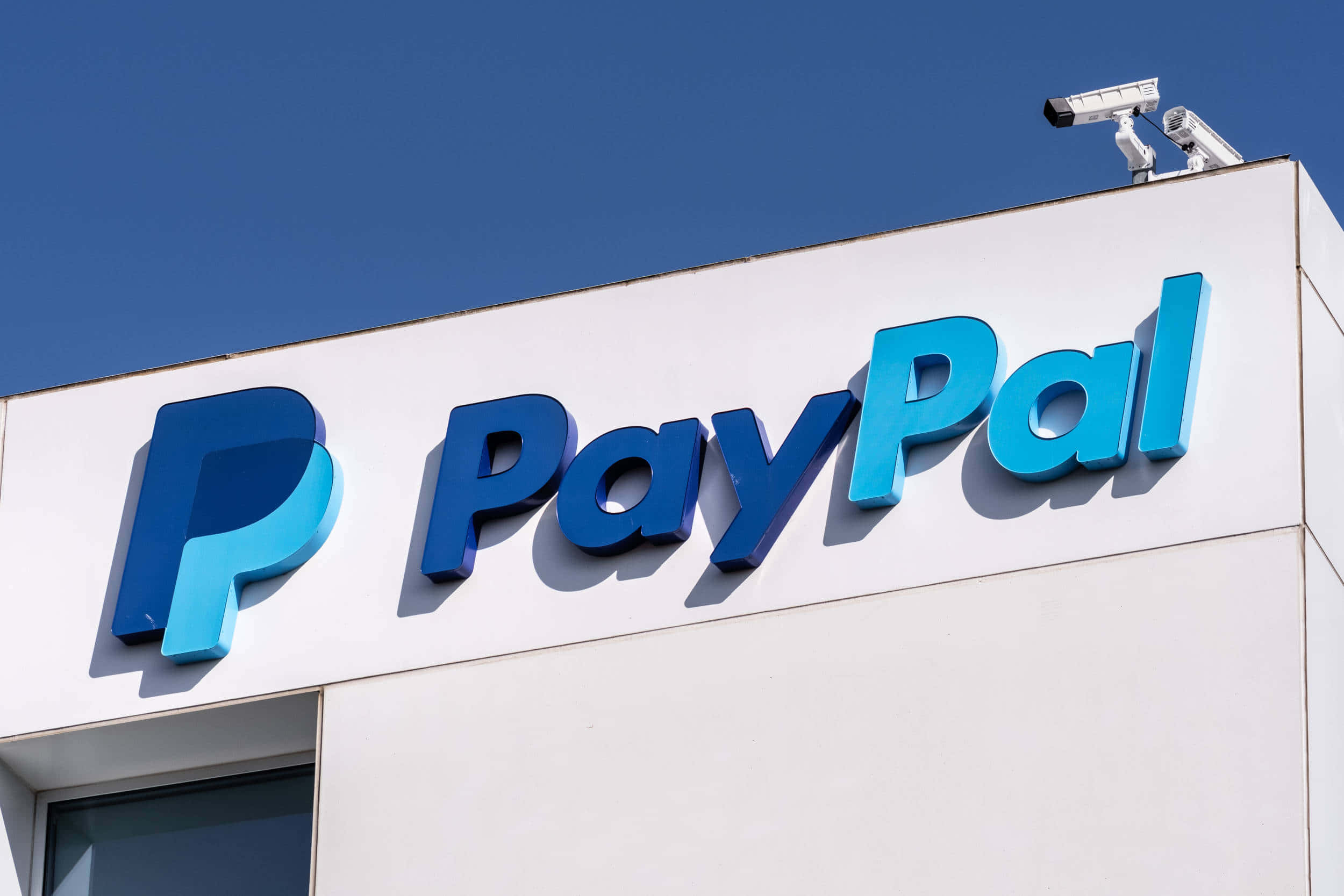 PayPal 桌布 壁紙 — 鮮明的藍色PayPal標誌安裝在白色建築物上，上方有藍天和保安攝影機。