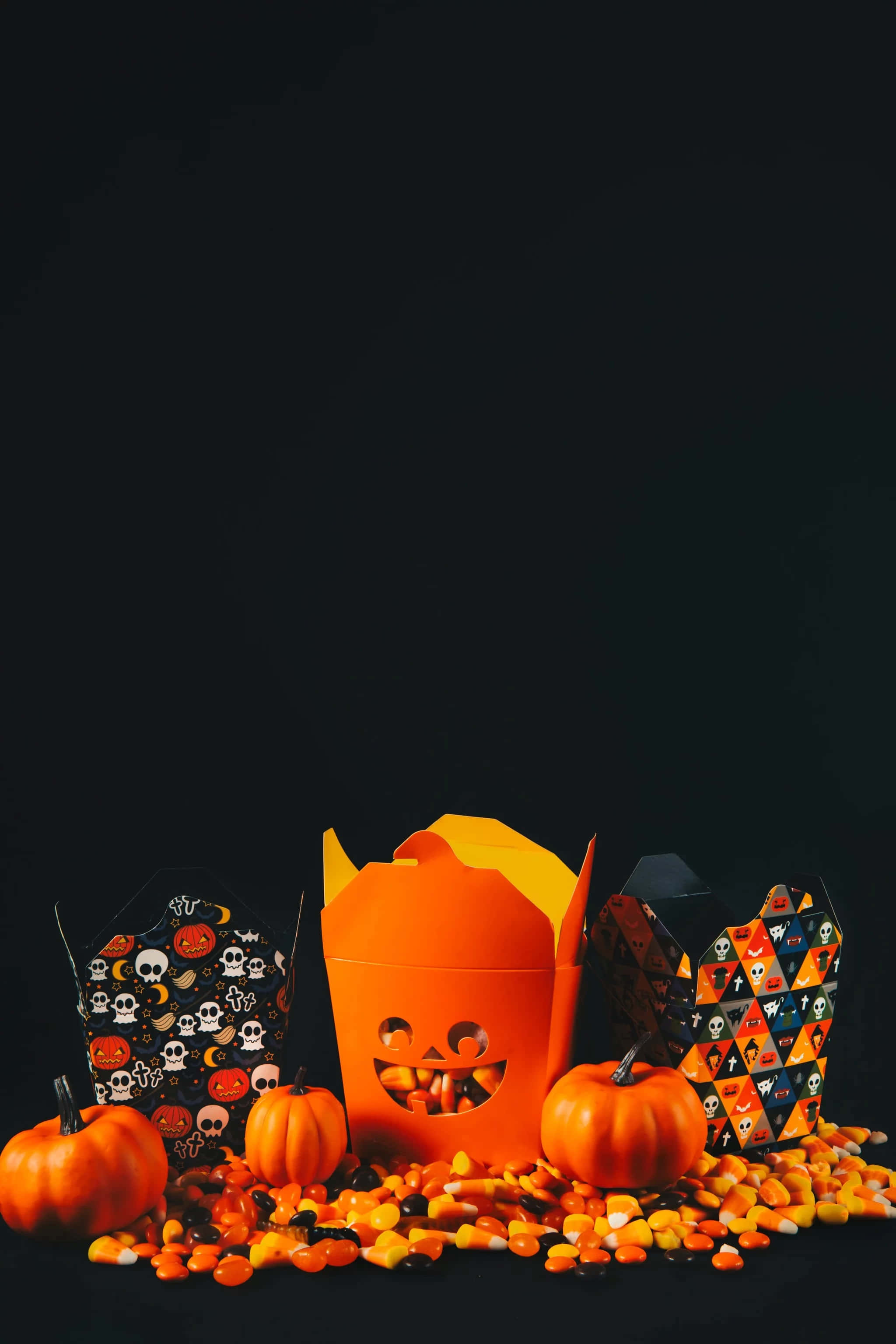 Halloween Phone 桌布 壁紙 — 鮮橙色南瓜形糖果盒和真正的南瓜在漆黑的背景上灑出糖果玉米，營造萬聖節氣氛。