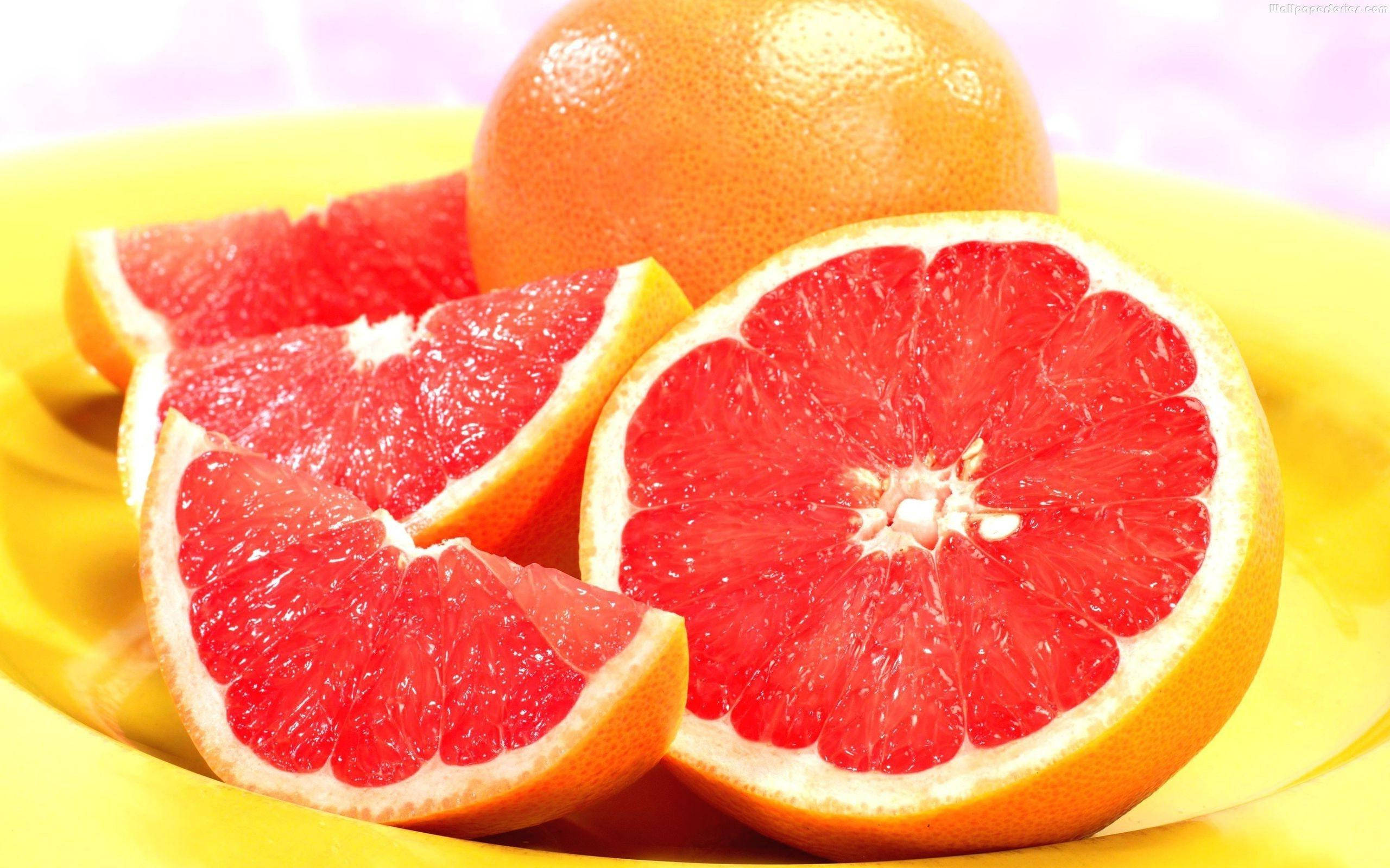 Grapefruit 桌布 壁紙 — 鮮紅的西柚切片和完整果實，在柔和的粉紅色背景下，於明亮的黃色盤子上閃閃發光。