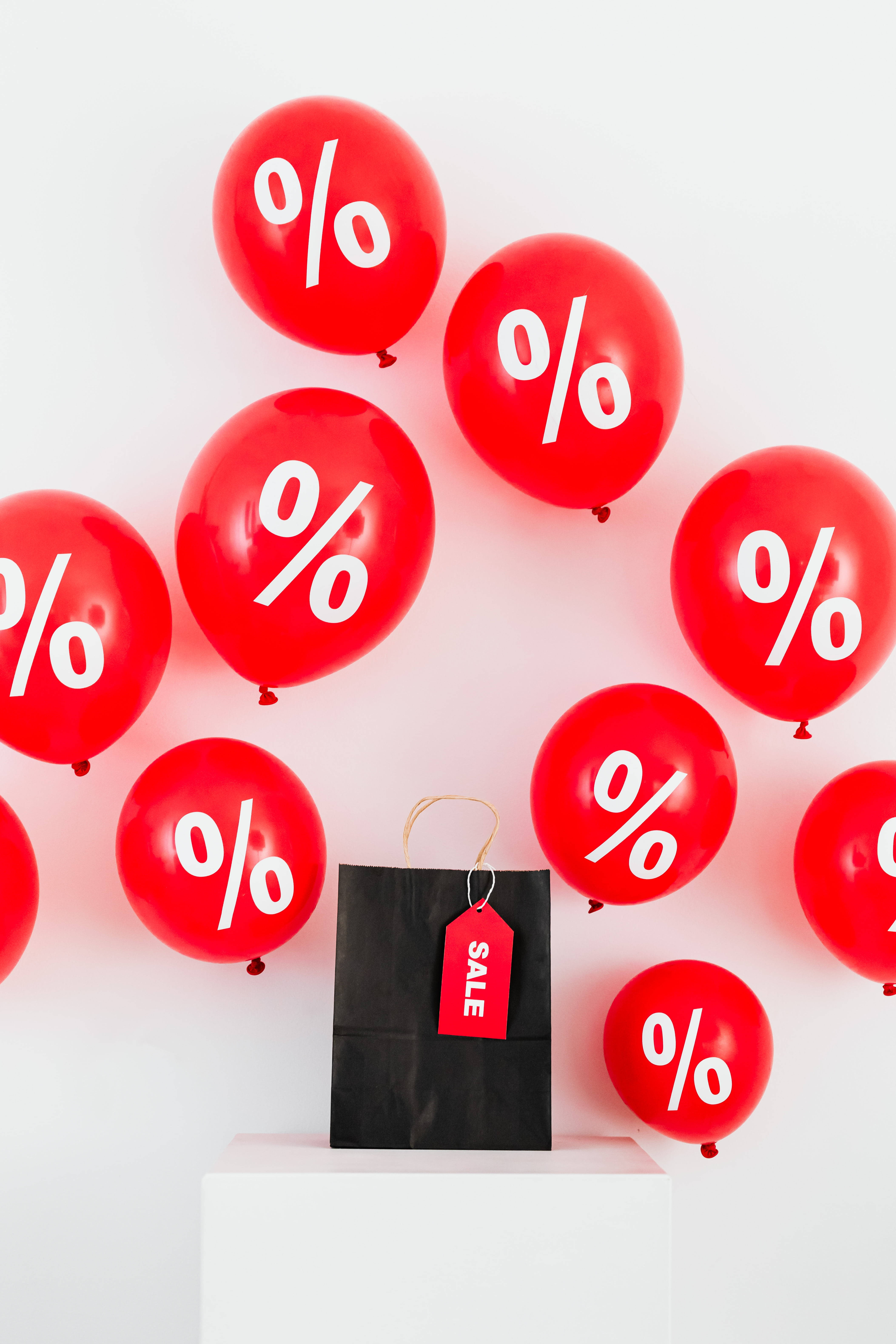 Sales 桌布 壁紙 — 鮮紅色氣球帶有白色百分比符號，漂浮在一個標有 sale 的黑色購物袋周圍，背景是乾淨的白色。