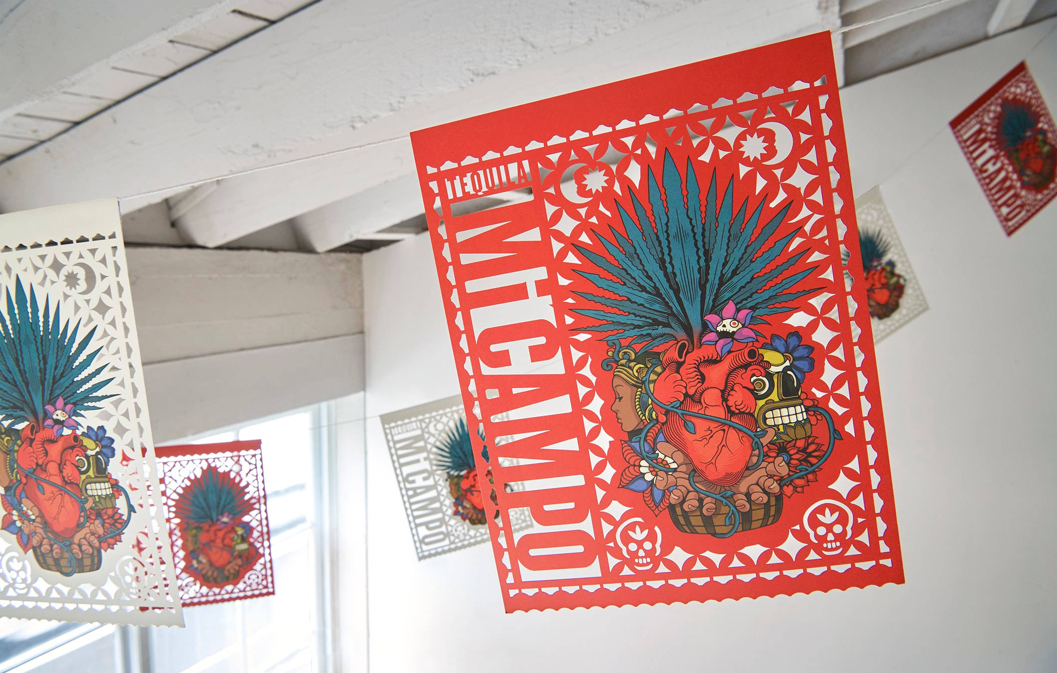 stickers 桌布 壁紙 — 鮮紅色的墨西哥剪紙旗幟，帶有精緻的骷髏、心形和龍舌蘭鏤空圖案，懸掛在明亮室內空間的白色天花板上。