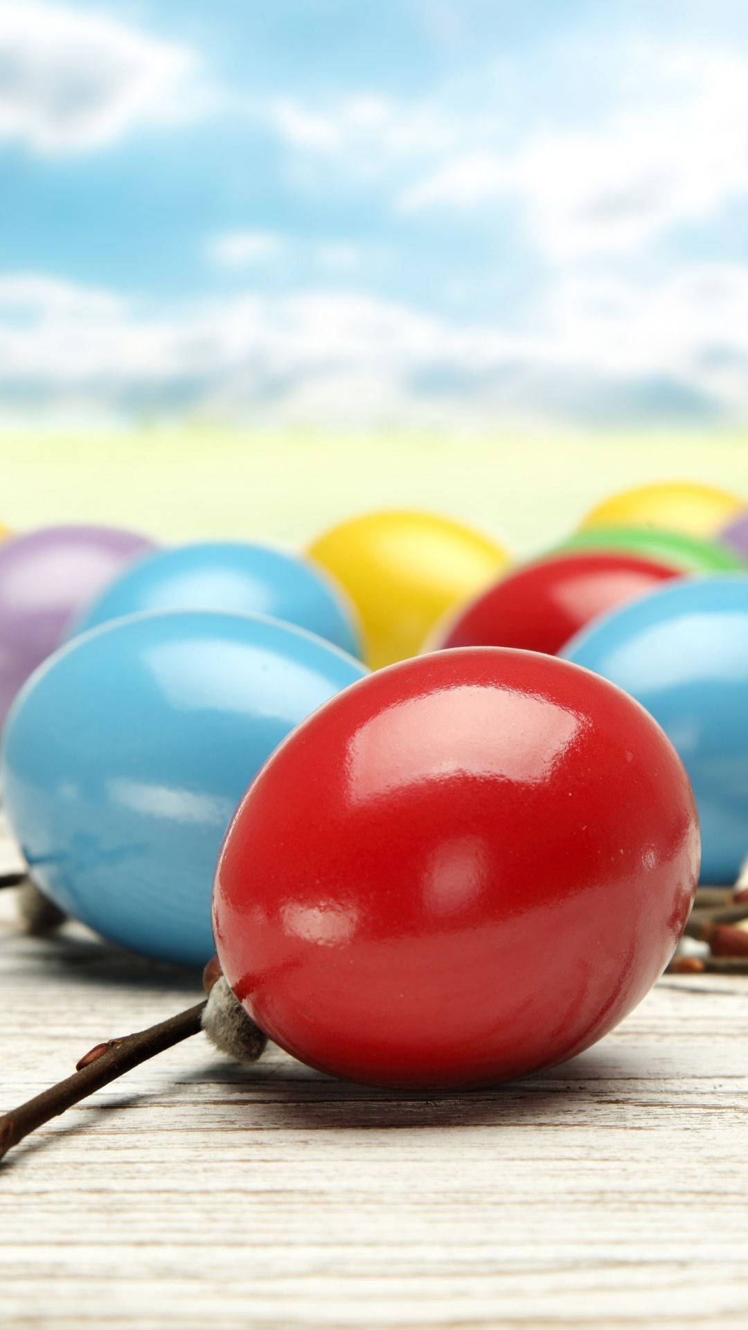 Easter iPhone 桌布 壁紙 — 鮮豔光澤的復活節彩蛋（紅色、藍色、黃色和紫色）放在風化的木頭上，背景是明亮的藍天和蓬鬆的白雲。