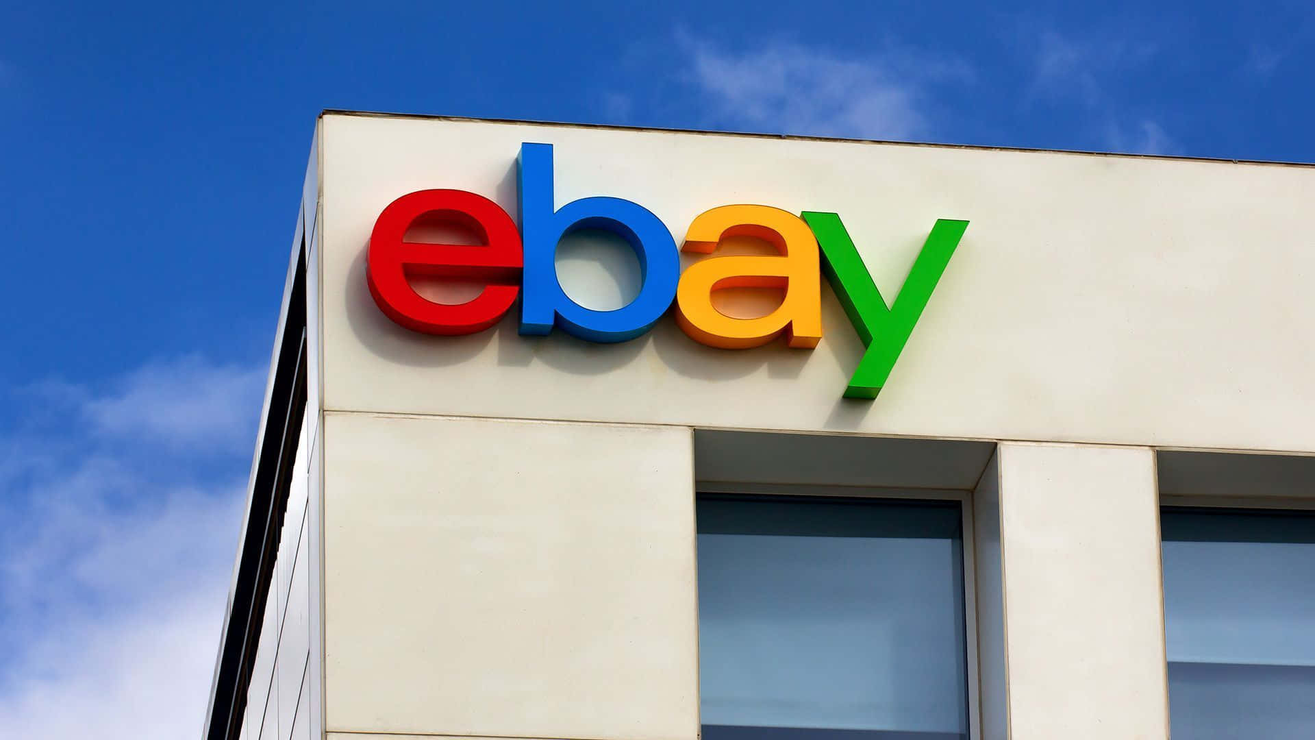 品牌 桌布 壁紙 — 鮮豔的eBay標誌，以紅、藍、黃、綠色呈現，安裝在現代建築上，背景是清澈的藍天和零星的白雲。