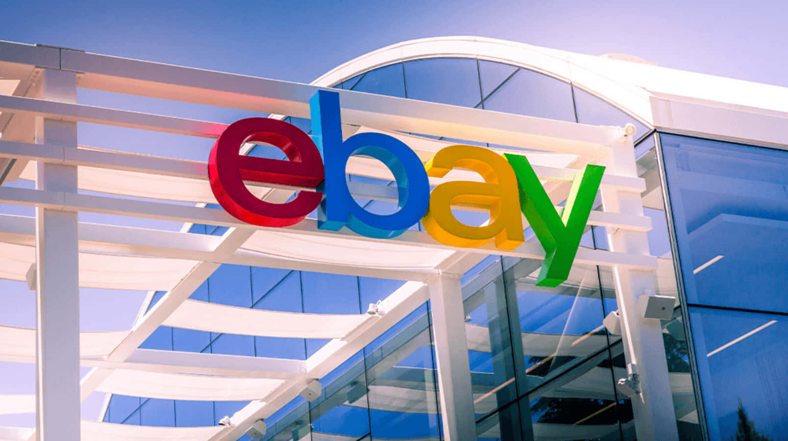 eBay 桌布 壁紙 — 鮮豔的eBay標誌，以紅、藍、黃、綠色呈現，安裝在現代玻璃與白色鋼結構建築外牆上，背景是清澈的藍天。