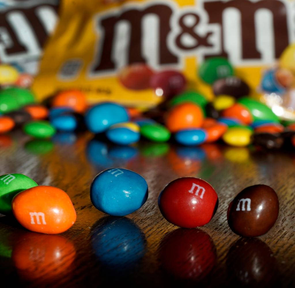 巧克力 桌布 壁紙 — 鮮豔的M&M's糖果在前景，後面有模糊的黃色袋子在反光的木質表面上。