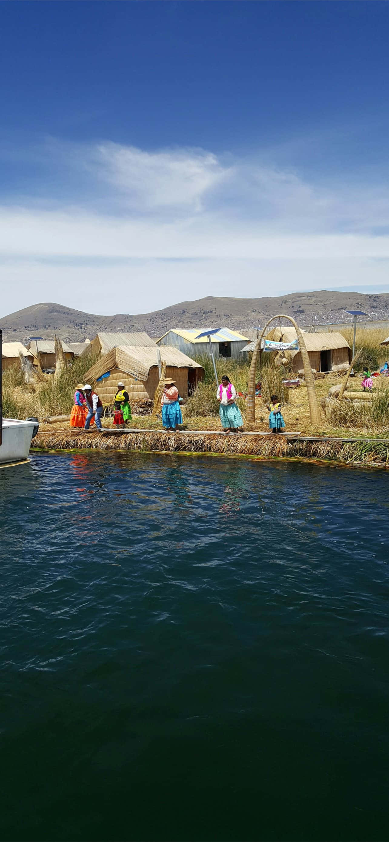 Islands 桌布 壁紙 — 鮮豔的傳統房屋建在漂浮的蘆葦島上，人們穿著色彩繽紛的服裝，靠近平靜的深色水域，上方是明亮的藍天。
