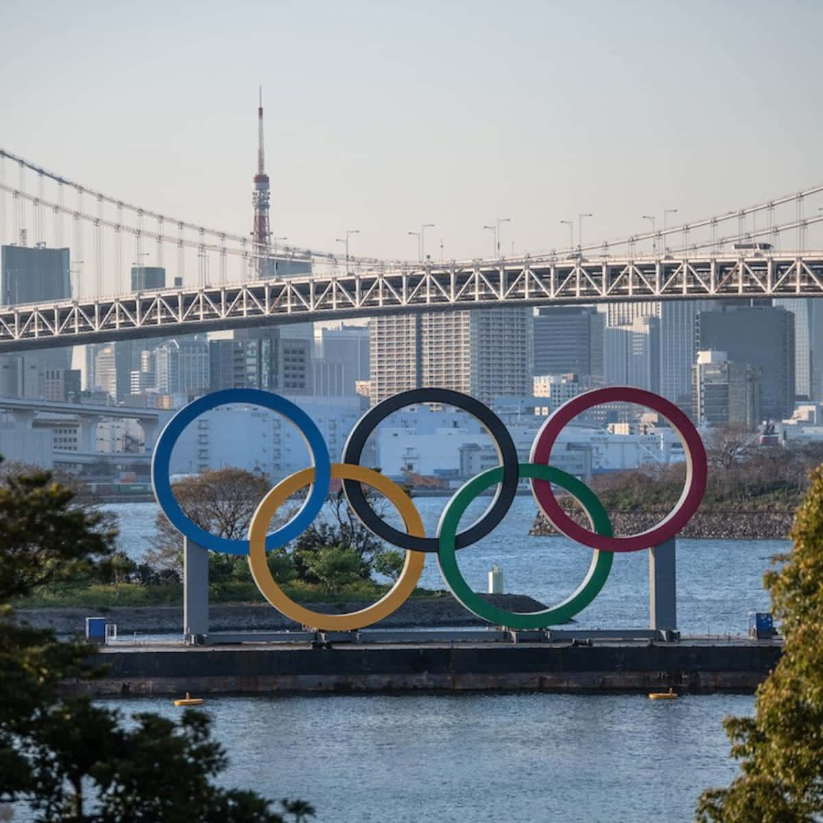 Olympics 桌布 壁紙 — 鮮豔的奧運五環矗立在水邊，東京鐵塔和彩虹橋在晴朗的白天高聳於後方。