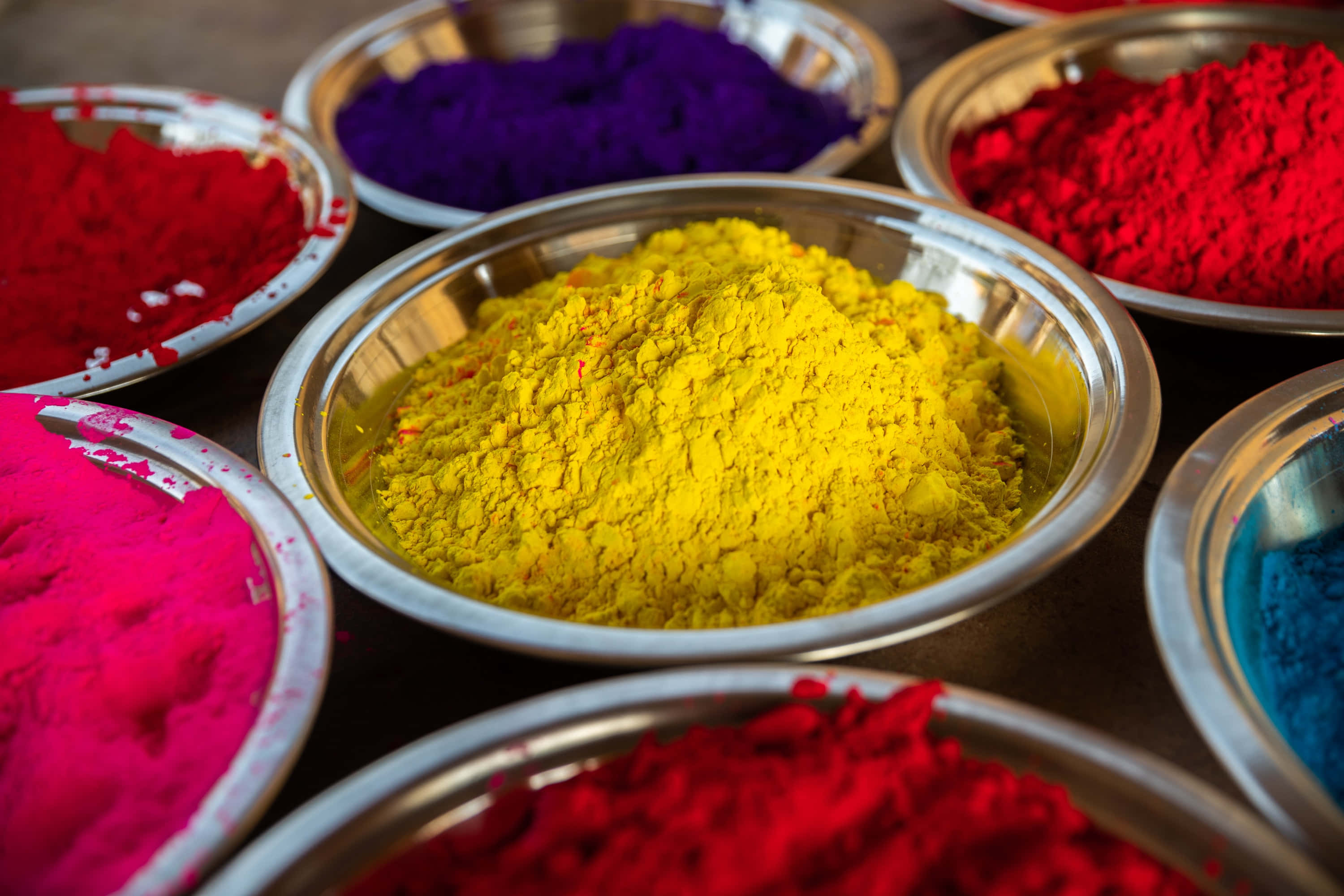 Holi 桌布 壁紙 — 鮮豔的彩色粉末放在閃亮的金屬碗中，以圓形圖案排列，帶有鮮明的紅色、紫色、黃色和藍色色調。