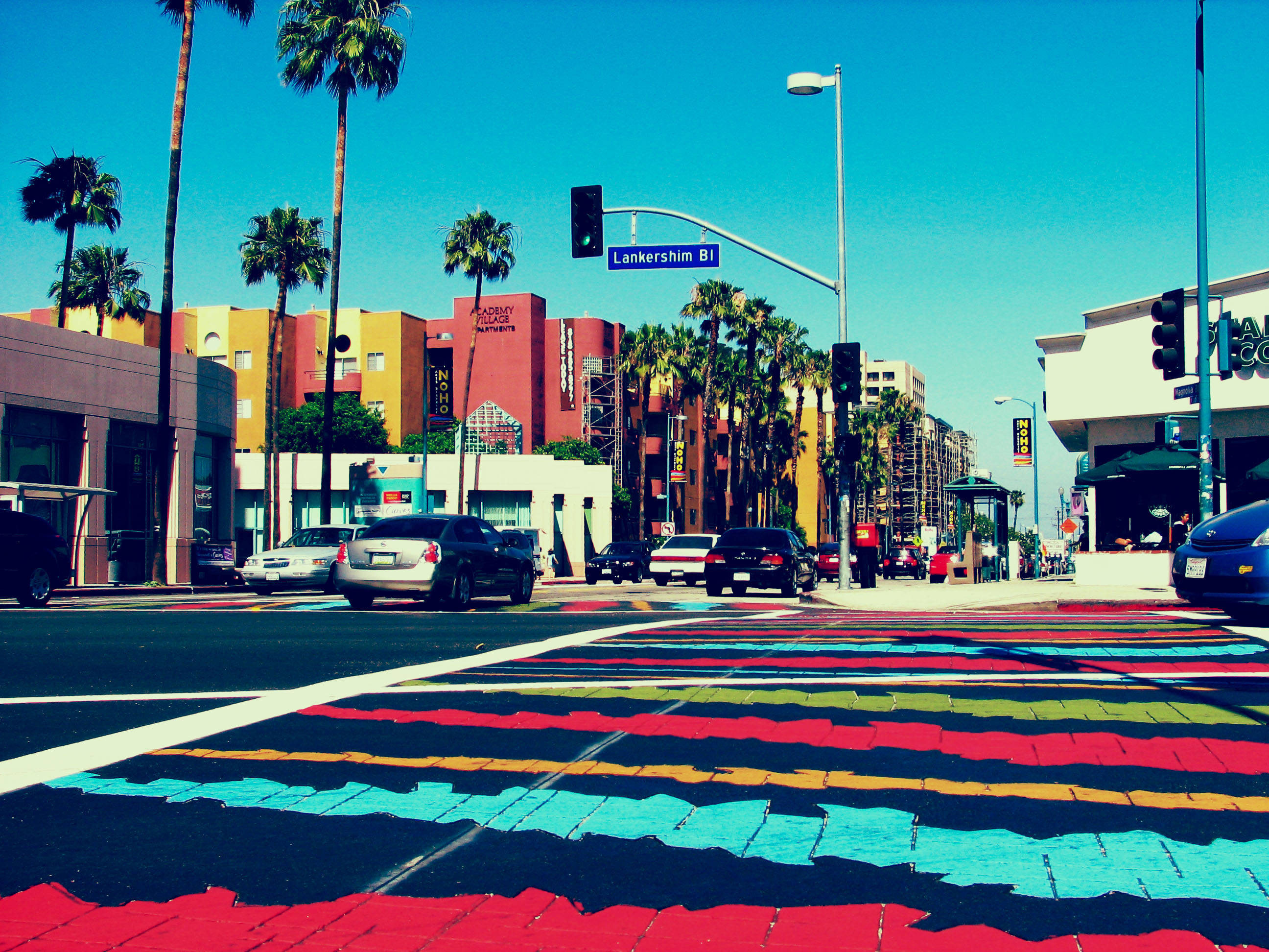hollywood street 桌布 壁紙 — 鮮豔的彩虹斑馬線延伸在一條陽光明媚的城市街道上，兩旁種有棕櫚樹和色彩繽紛的建築物，在清澈的藍天下。