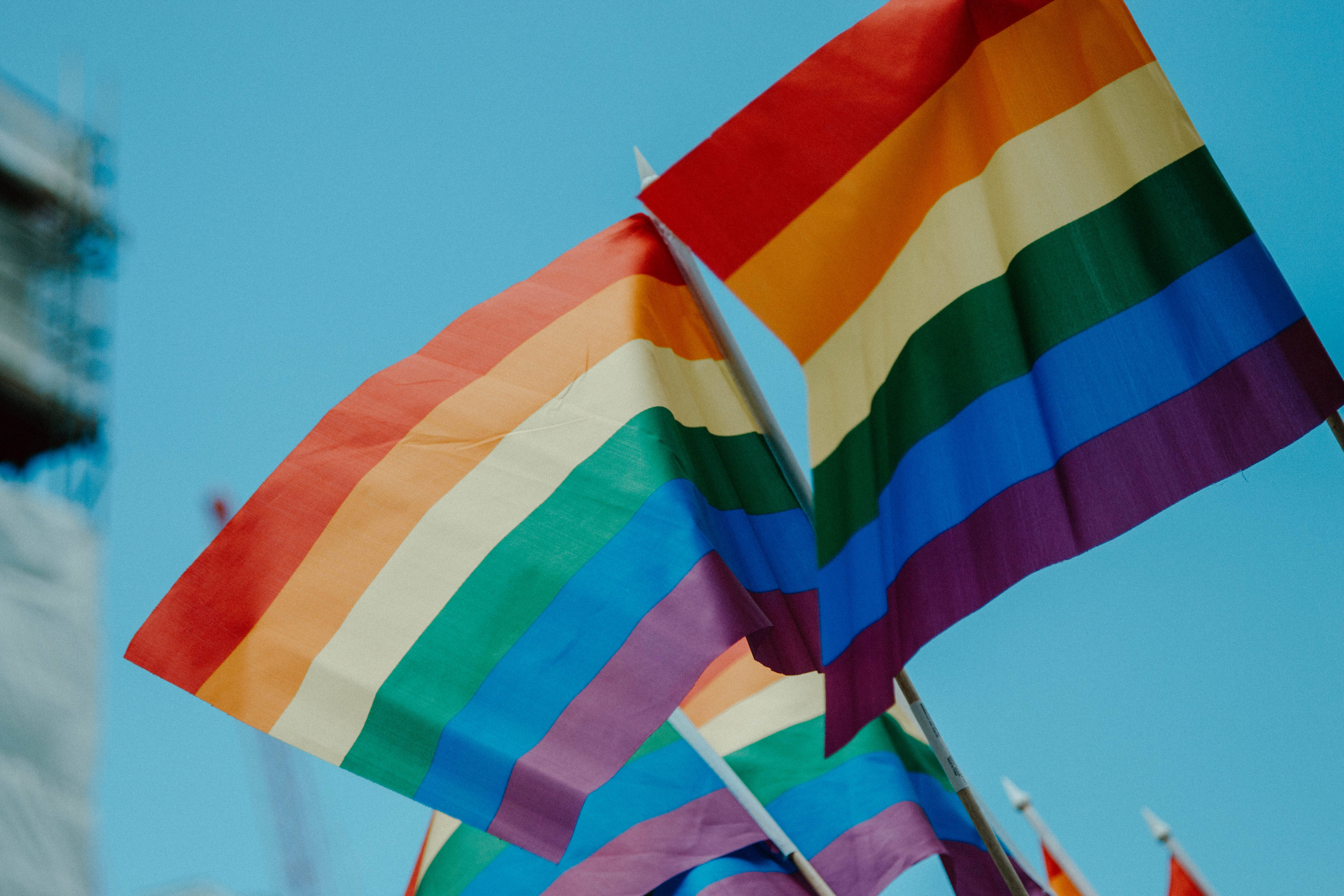 LGBT 桌布 壁紙 — 鮮豔的彩虹旗在清澈的藍天下飄揚，背景有一座模糊的建築物。