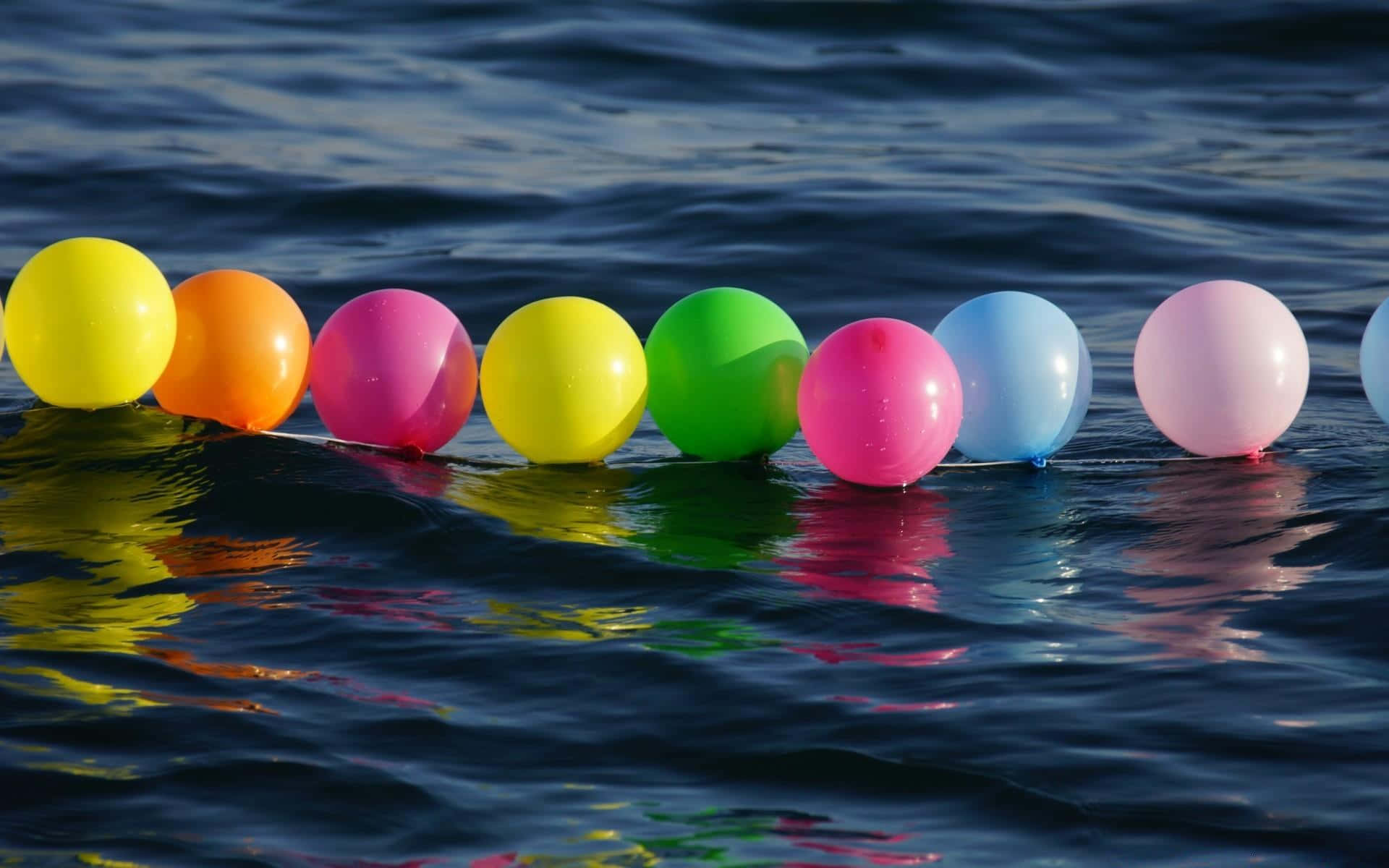 Balloon 桌布 壁紙 — 鮮豔的彩虹氣球漂浮在漣漪的深藍色水面上，反射出它們明亮的色彩。