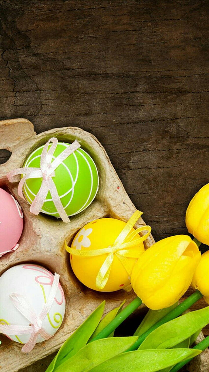 Easter Phone 桌布 壁紙 — 鮮豔的復活節彩蛋和黃色鬱金香放在深色木質表面上的質樸紙板托盤中。
