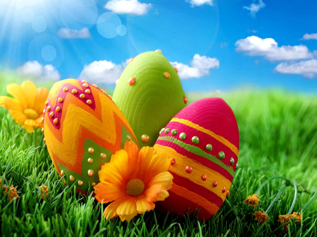 Cute Happy Easter 桌布 壁紙 — 鮮豔的手繪復活節彩蛋帶有金屬飾釘，放置在茂盛的綠草中，旁邊是鮮豔的橙色花朵，在晴朗的藍天下。