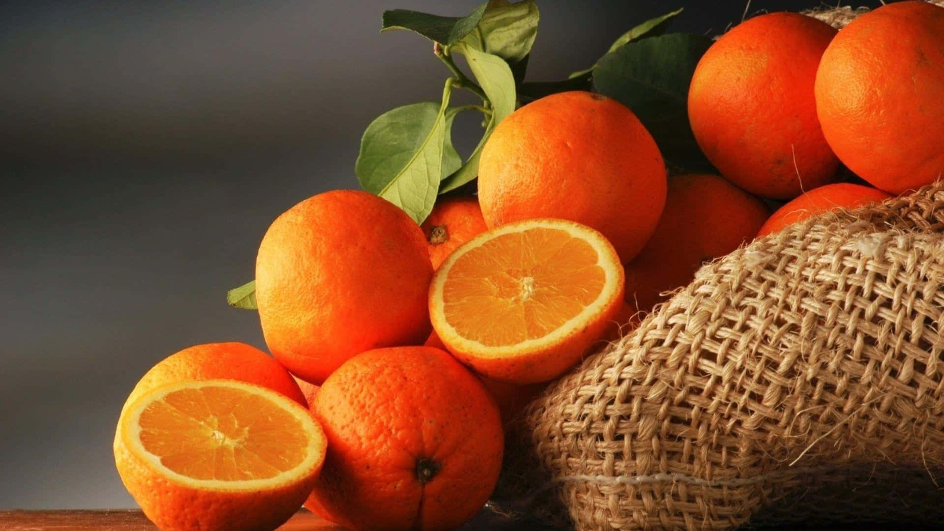 Orange Fruit 桌布 壁紙 — 鮮豔的柳橙、綠葉和切半的果實，放在深色背景旁的粗糙麻布袋上。
