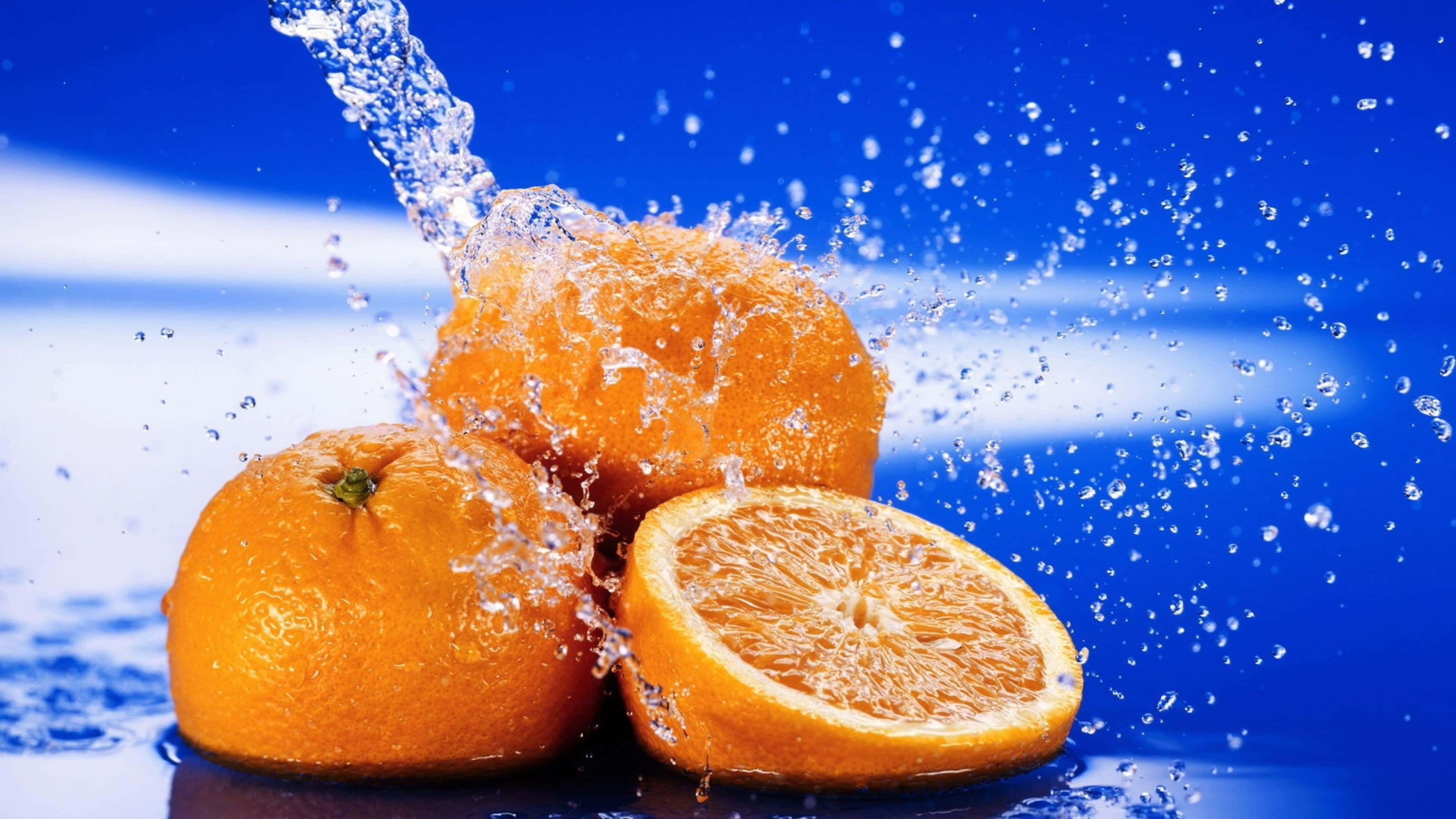 Orange Fruit 桌布 壁紙 — 鮮豔的橙子在強烈的藍色背景下與水一起爆裂，形成動態的水滴和多汁的新鮮感。