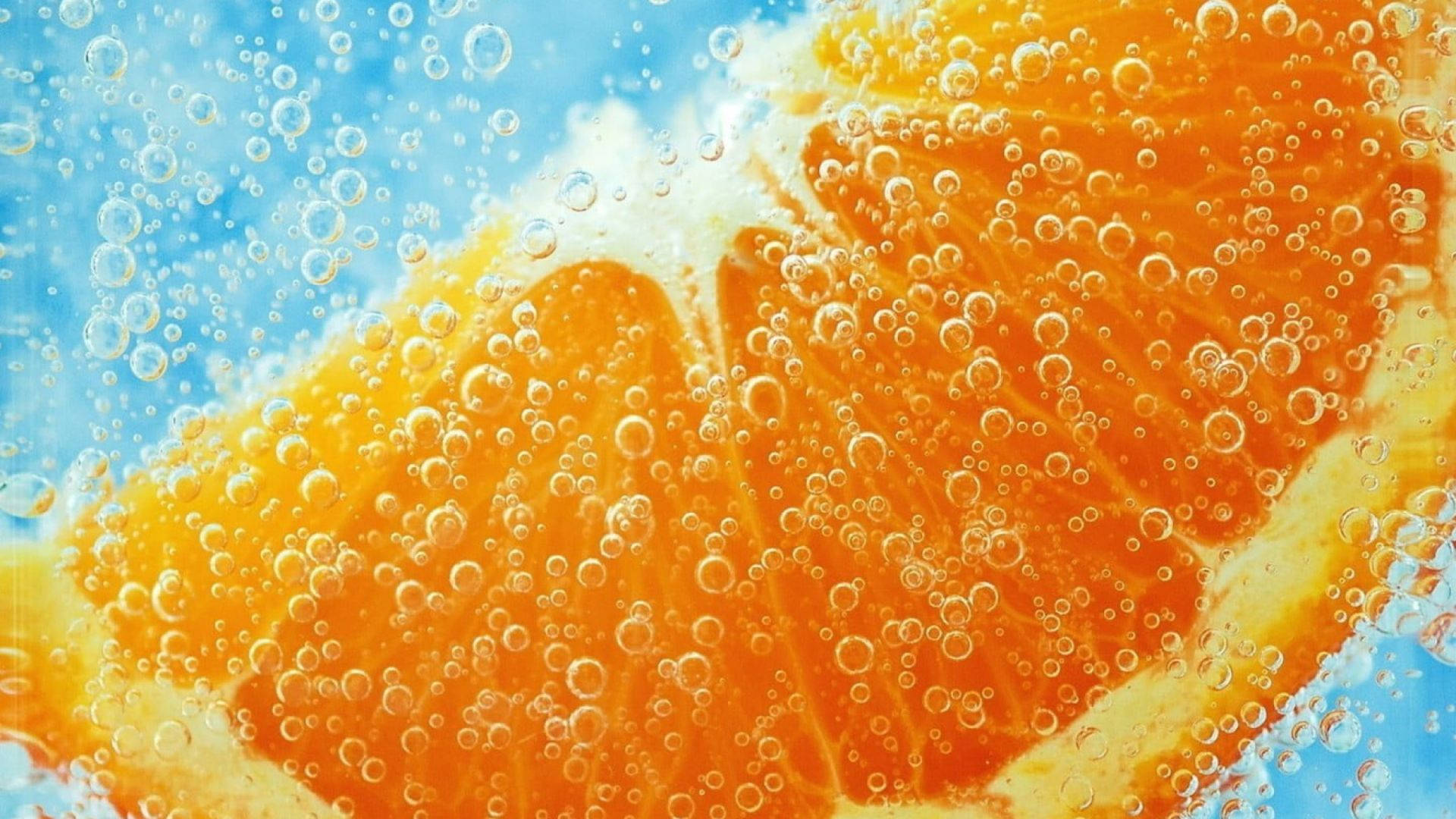 Orange Fruit 桌布 壁紙 — 鮮豔的橙片浸在氣泡水中，氣泡在明亮的藍色背景上升起。