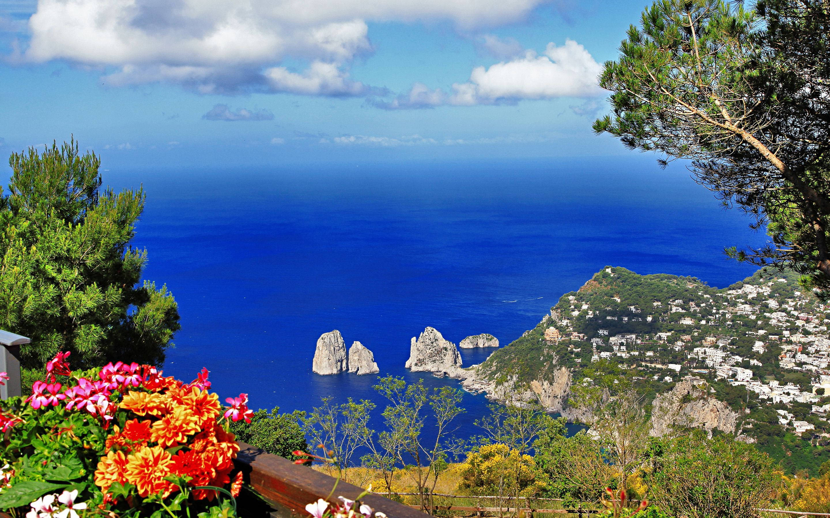 Capri 桌布 壁紙 — 鮮豔的橙色和粉紅色花朵在前景盛開，俯瞰深藍色的大海、標誌性的法拉廖尼岩石和明亮天空下的沿海村莊。