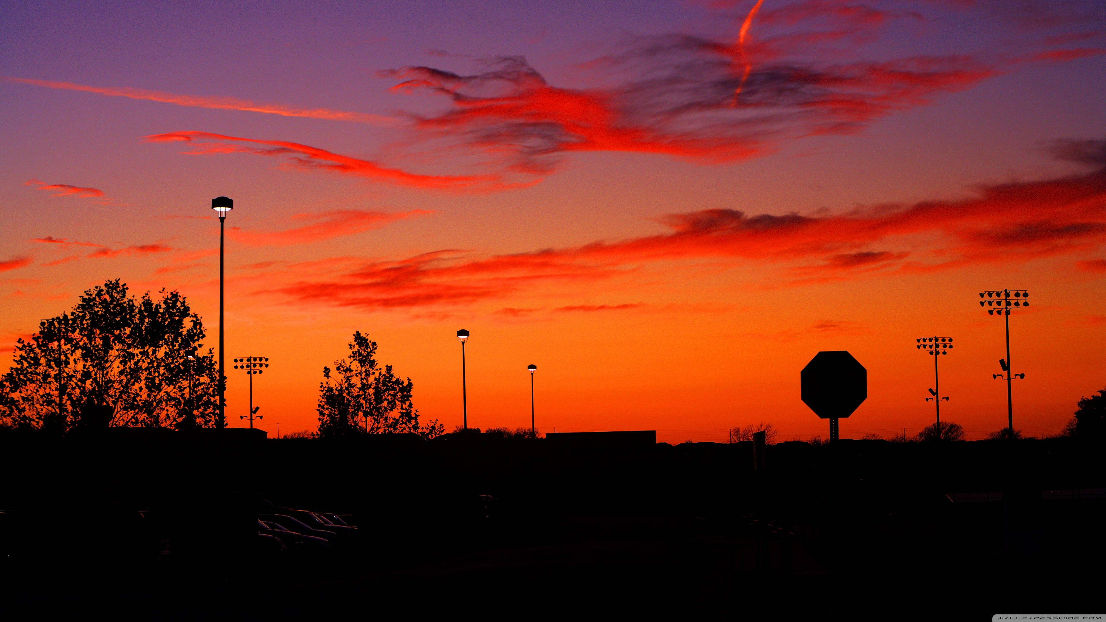 Tumblr Sunset Desktop 桌布 壁紙 — 鮮豔的橙色和紫色日落天空勾勒出體育場燈光、樹木和停車標誌的剪影，背景是黑暗的前景。
