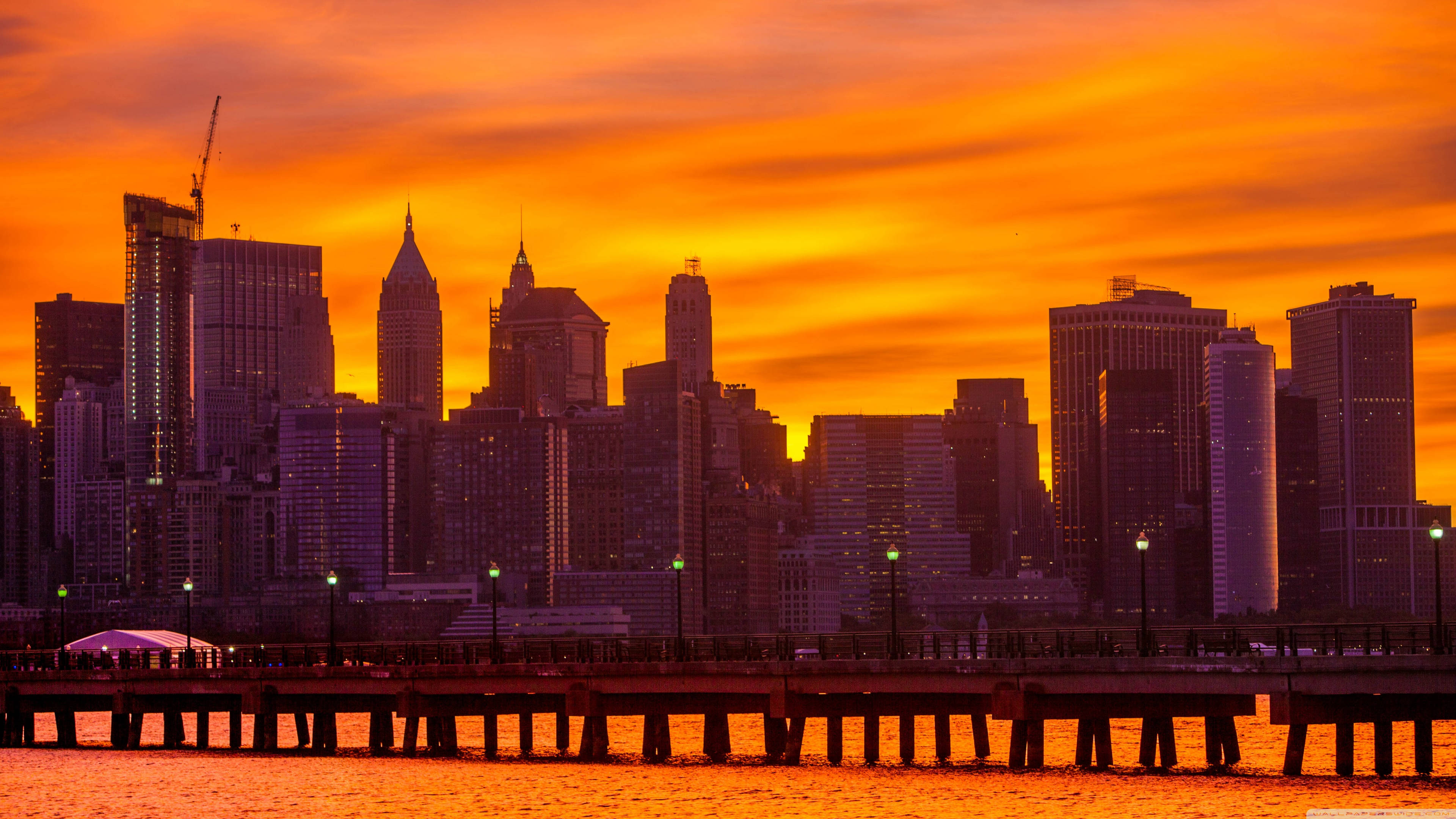 4K New York City 桌布 壁紙 — 鮮豔的橙色夕陽將紐約市天際線沐浴在溫暖的光芒中，剪影般的摩天大樓矗立在海濱碼頭之上。