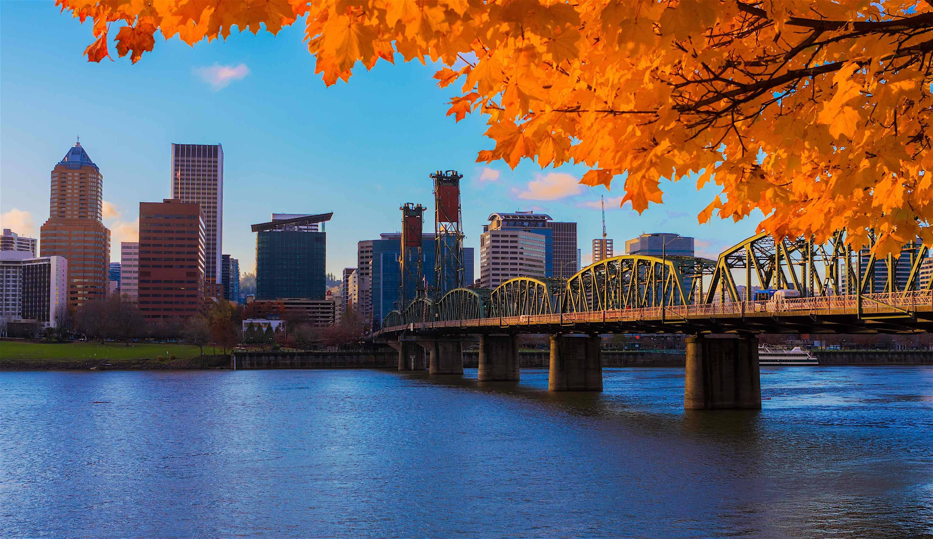 Portland 桌布 壁紙 — 鮮豔的橙色秋葉框住了城市天際線，一座黃色橋樑橫跨平靜的藍色河流，天空晴朗。