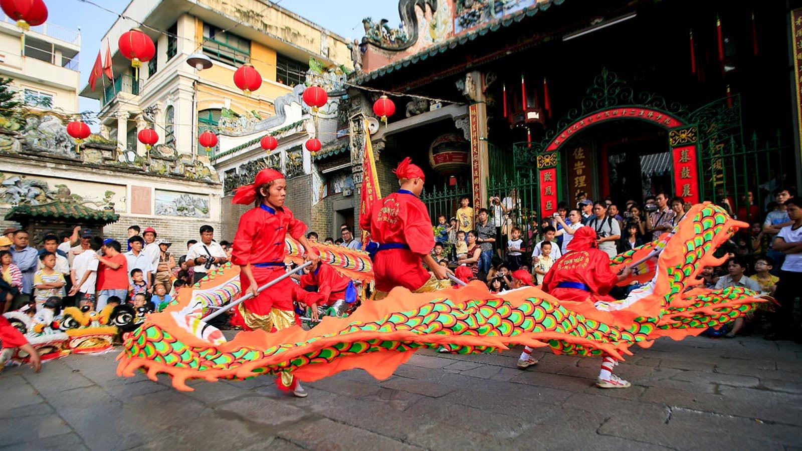 Ho Chi Minh City 桌布 壁紙 — 鮮豔的橙色舞龍者穿著紅色服裝，在裝飾著紅色燈籠的寺廟前進行傳統舞蹈，人群圍觀。