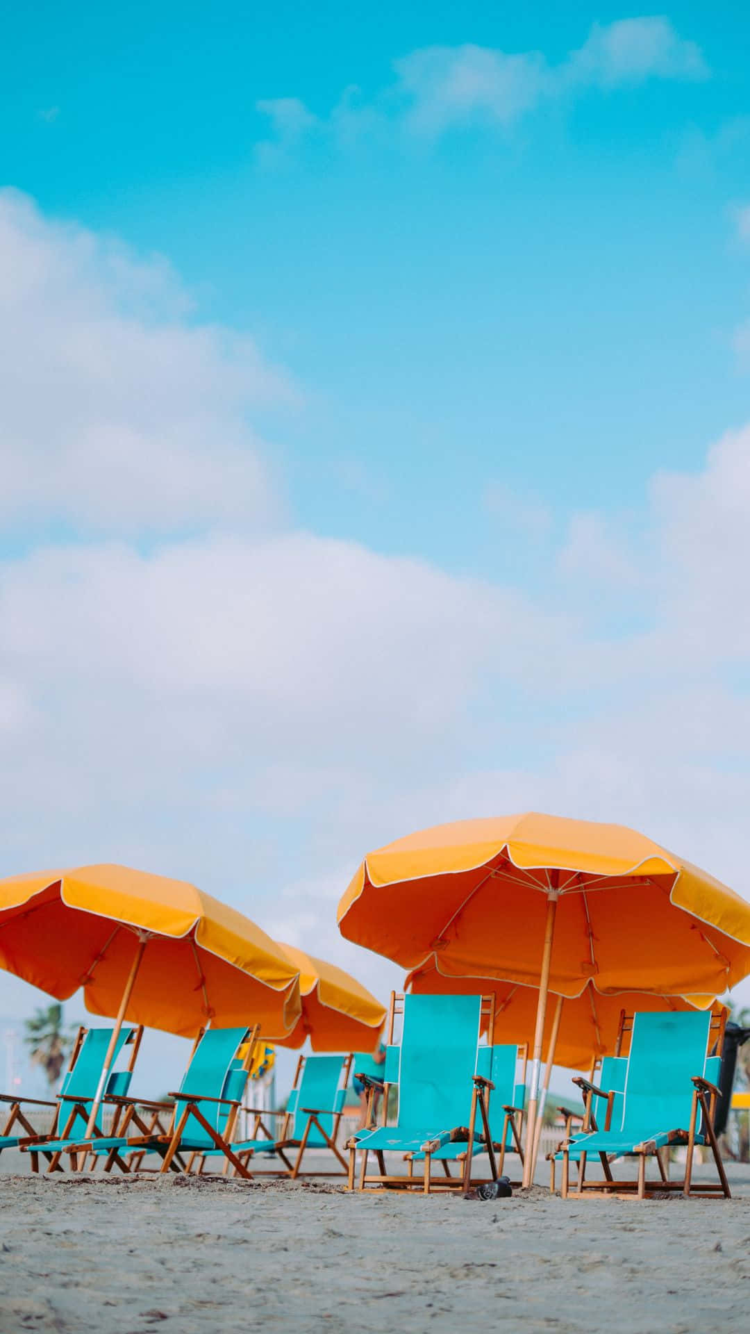 Android Summer 桌布 壁紙 — 鮮豔的橙色遮陽傘和藍綠色椅子點綴在陽光明媚的沙灘上，天空湛藍，白雲蓬鬆。