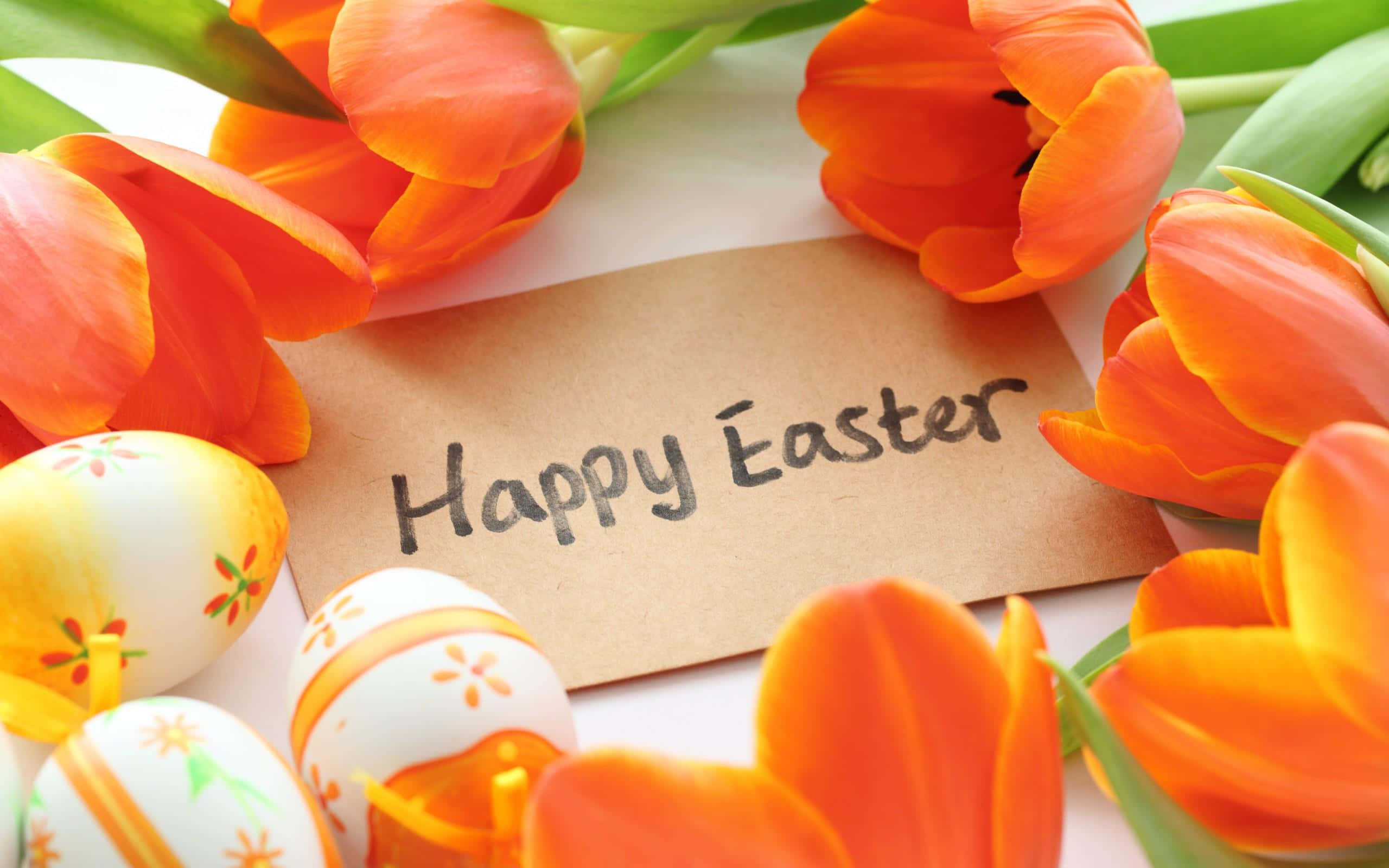 Cute Happy Easter 桌布 壁紙 — 鮮豔的橙色鬱金香圍繞著一張手寫的「復活節快樂」便條，旁邊是白色表面上色彩繽紛的裝飾蛋。
