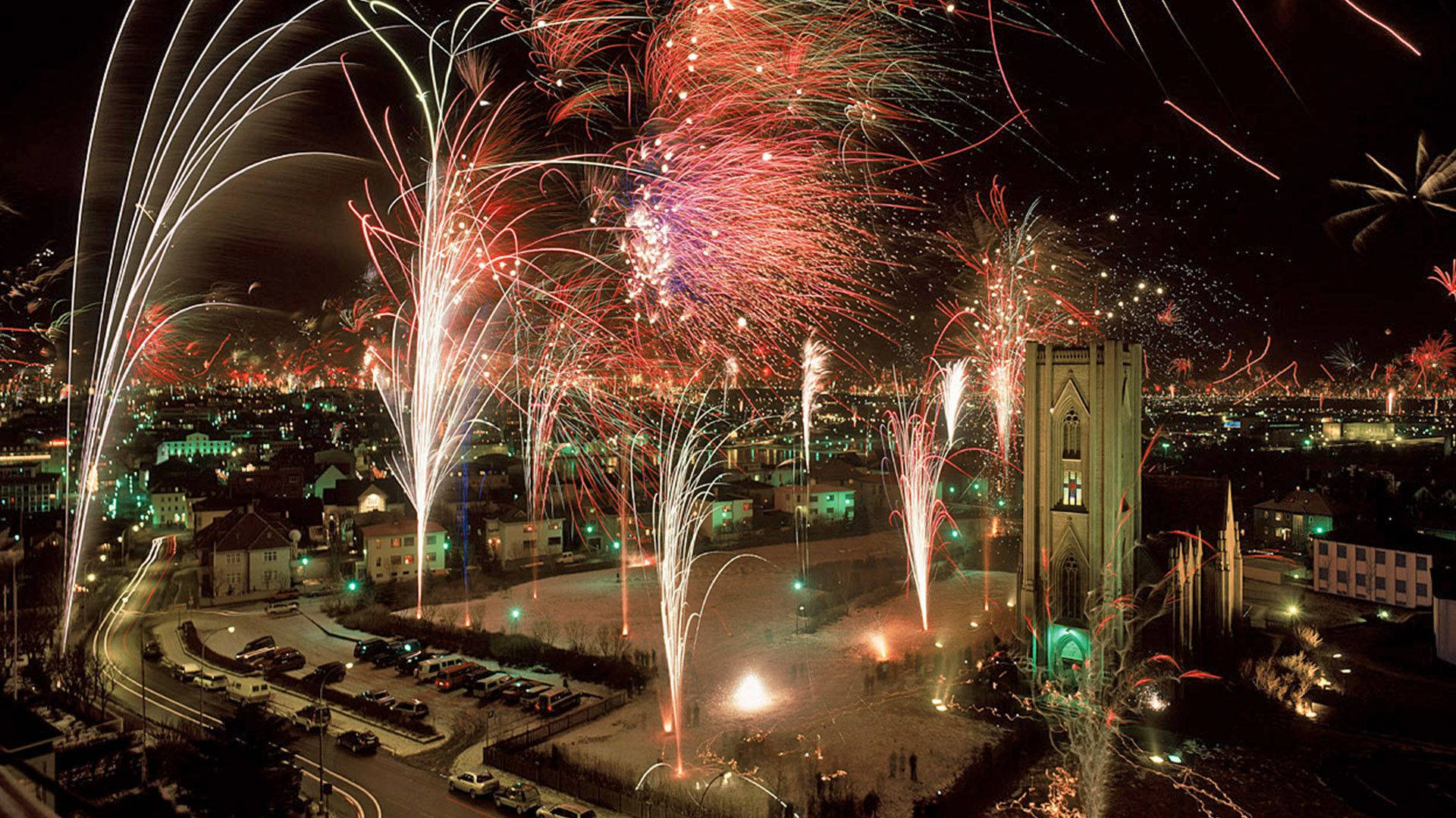 Fireworks 桌布 壁紙 — 鮮豔的煙火在夜間城市景觀上空爆炸，照亮了教堂塔樓和下面的街道。