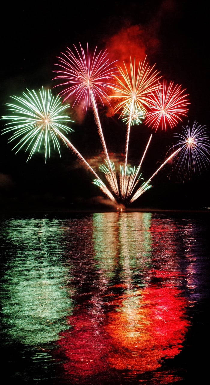 Fireworks 桌布 壁紙 — 鮮豔的煙火在水面上爆炸，在下方黑暗的水面上投射出色彩繽紛的倒影。