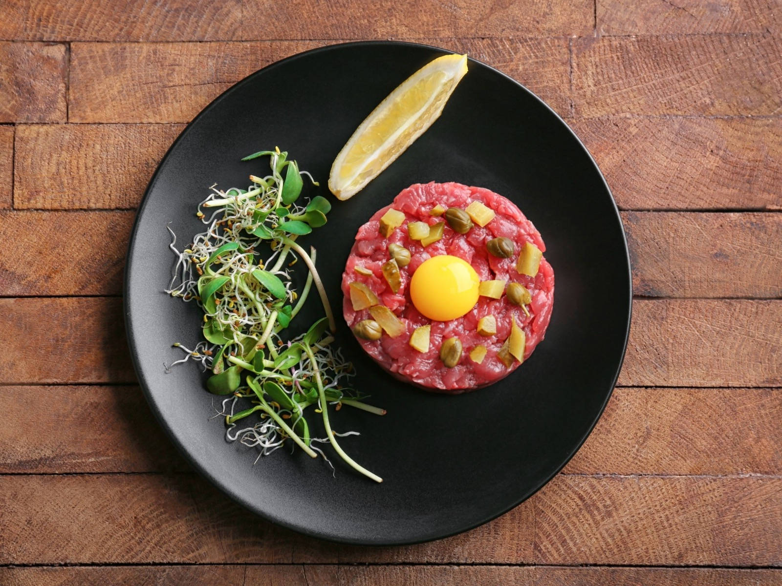meat 桌布 壁紙 — 鮮豔的生牛肉他他，上面放有生蛋黃、酸豆和醃菜，盛在黑色盤子上，配上微型蔬菜和檸檬角，置於鄉村風木板上。