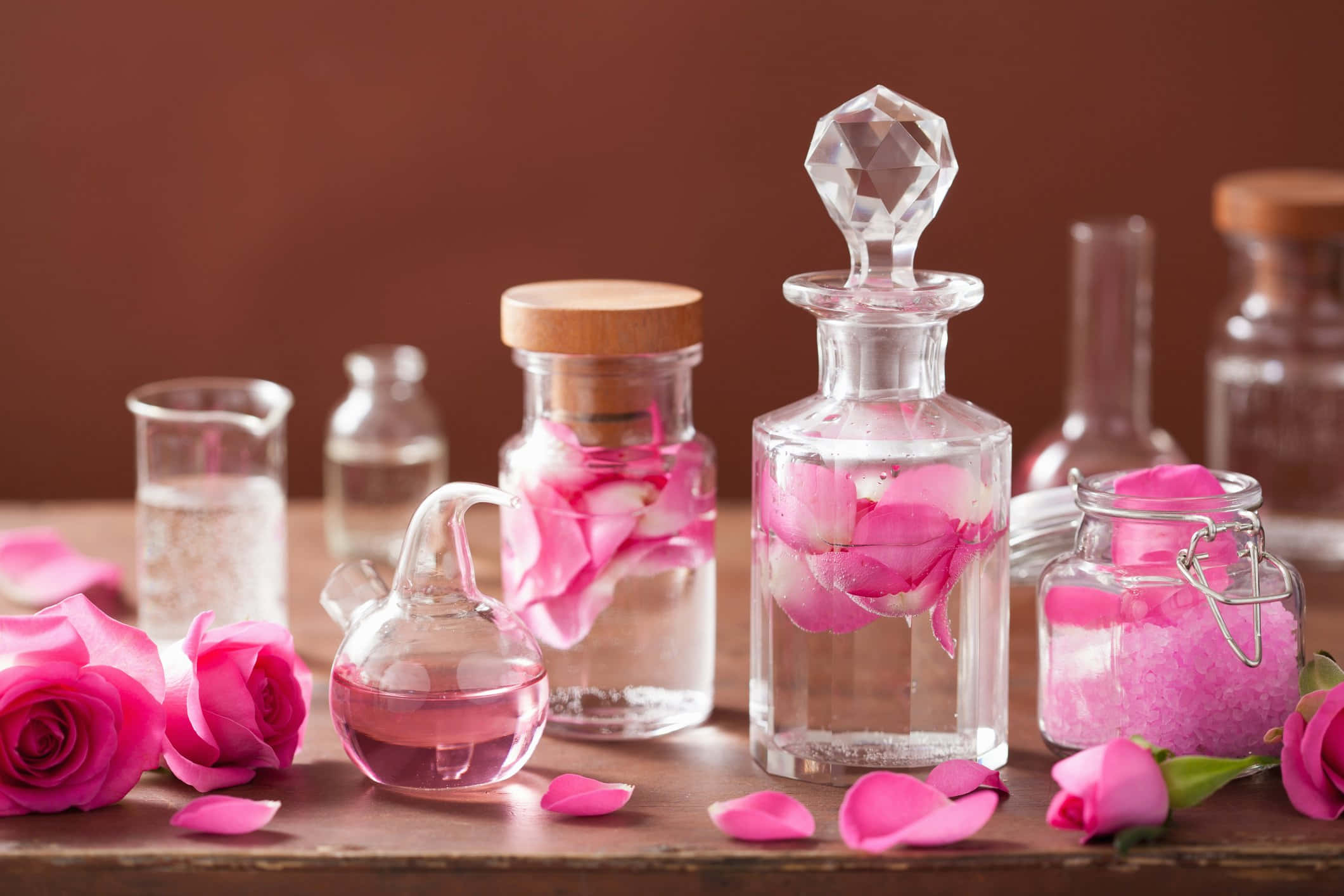 Aromatherapy 桌布 壁紙 — 鮮豔的粉紅玫瑰花瓣漂浮在裝滿清澈液體的玻璃瓶中，放在木質表面上，背景是溫暖的棕色。