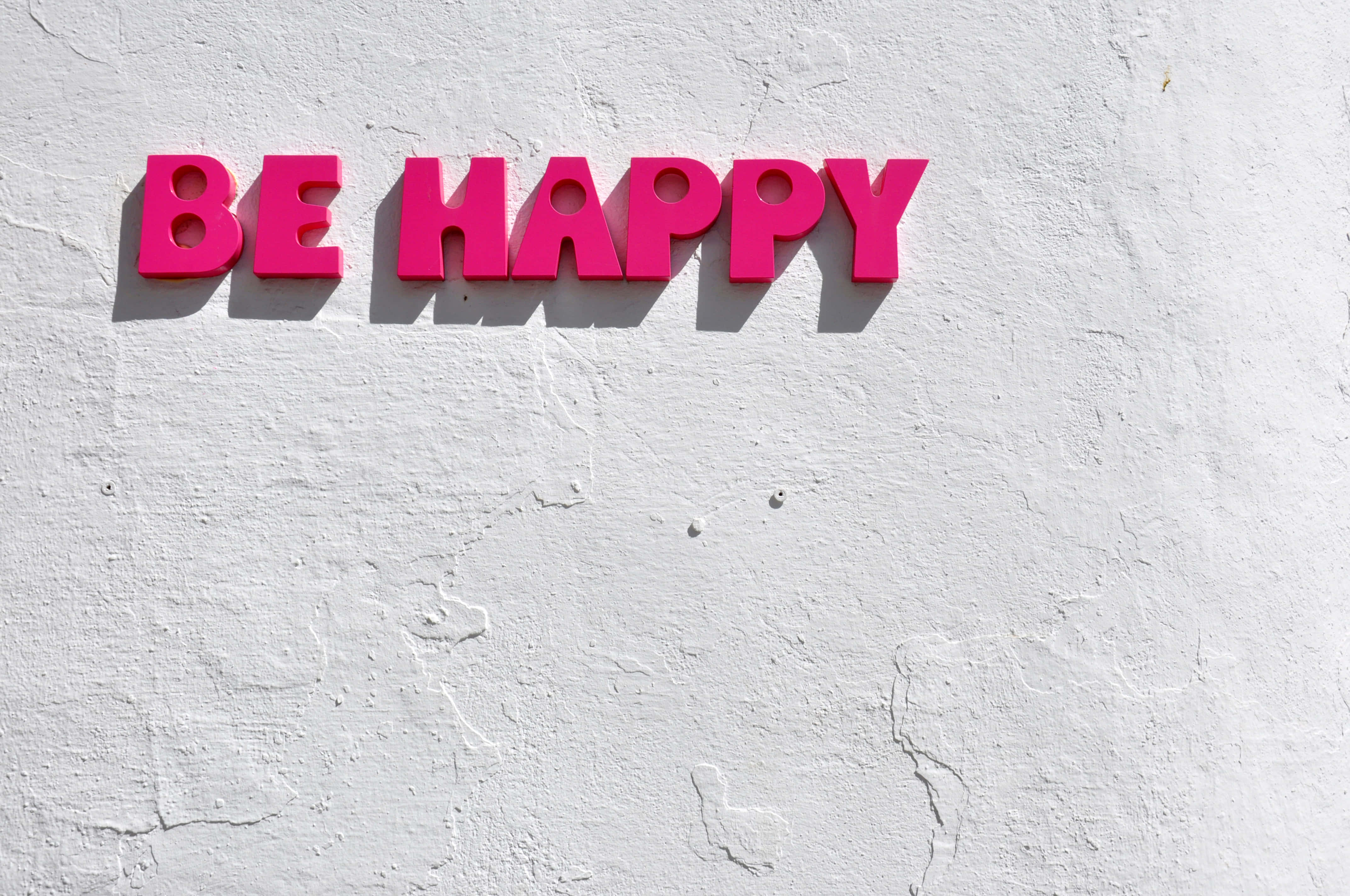 Happy Desktop 桌布 壁紙 — 鮮豔的粉紅色3D字母拼出BE HAPPY，在明亮的日光下，在粗糙的白色紋理牆上投下清晰的陰影。