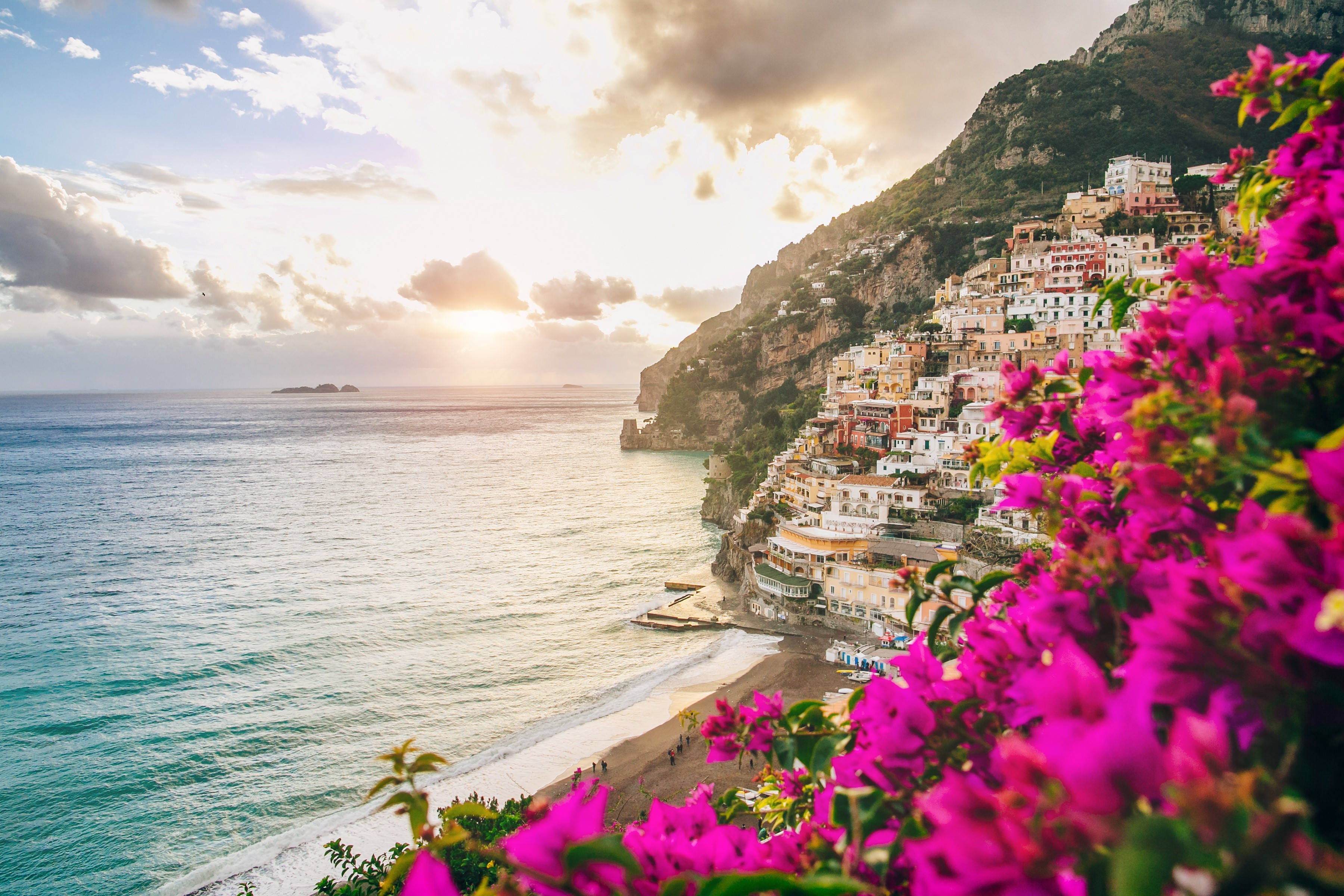 Amalfi Coast 桌布 壁紙 — 鮮豔的粉紅色九重葛環繞著一個陽光明媚的海岸村莊，村莊依附在懸崖上，俯瞰著日落時分的碧綠海水。