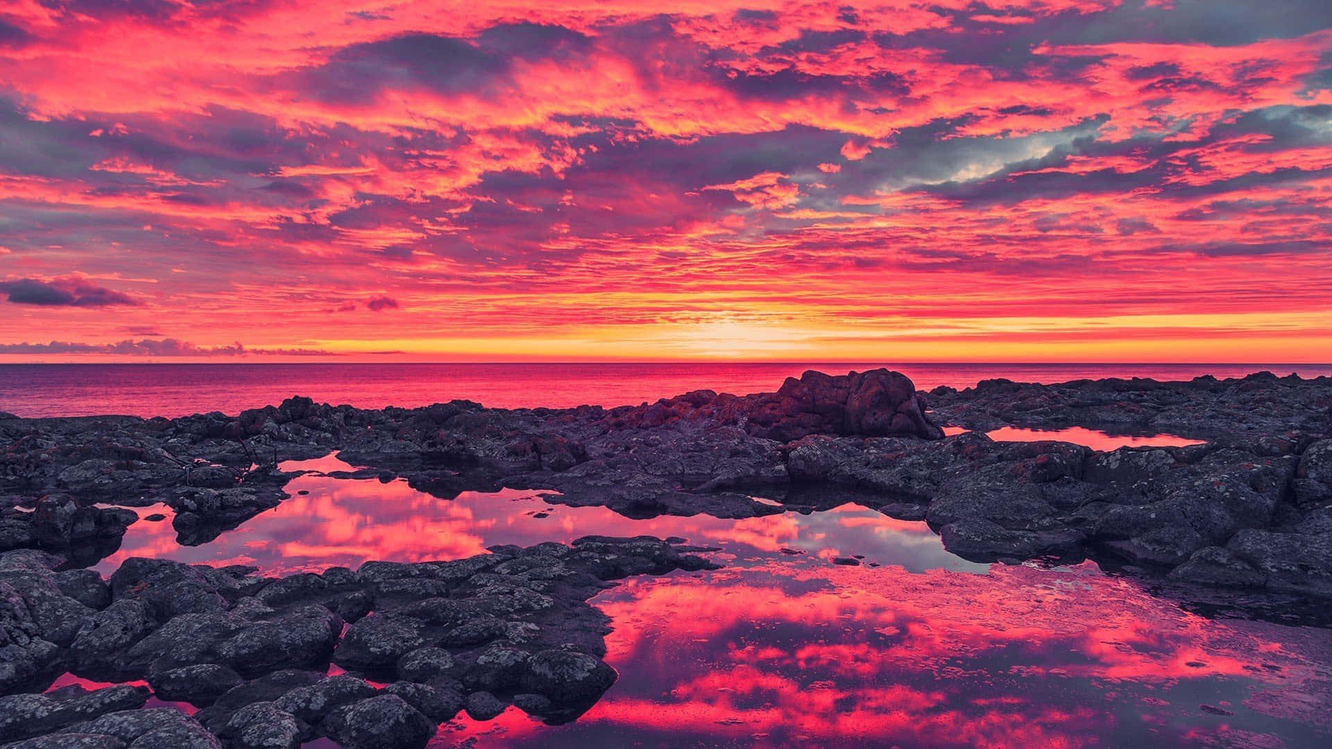 Pink Sky 桌布 壁紙 — 鮮豔的粉紅色和橙色日落雲彩倒映在海洋邊緣黑色火山岩之間的潮汐池中。