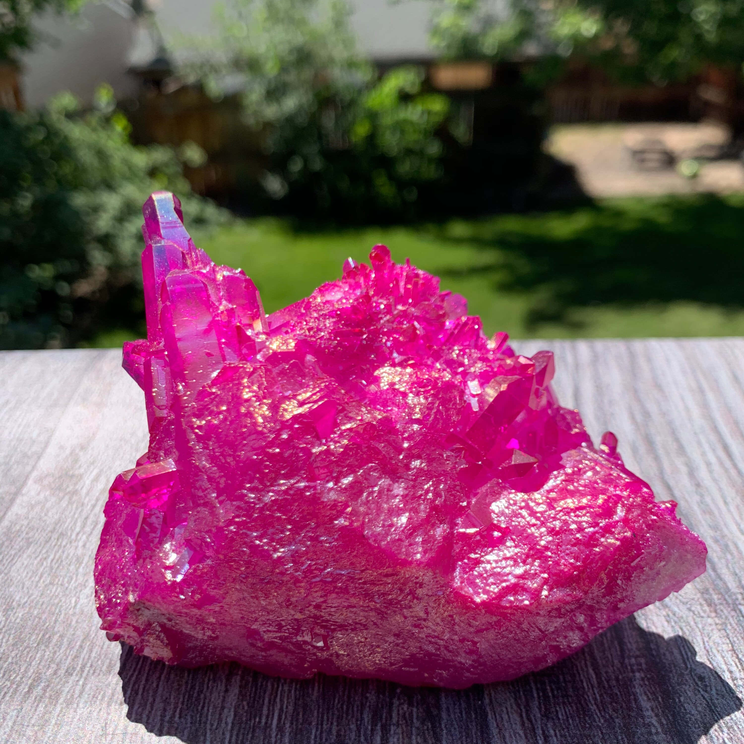 Pink Crystal Quartz 桌布 壁紙 — 鮮豔的粉紅色水晶簇在明亮的陽光下閃耀，放在木桌上，背景是模糊的綠色花園。
