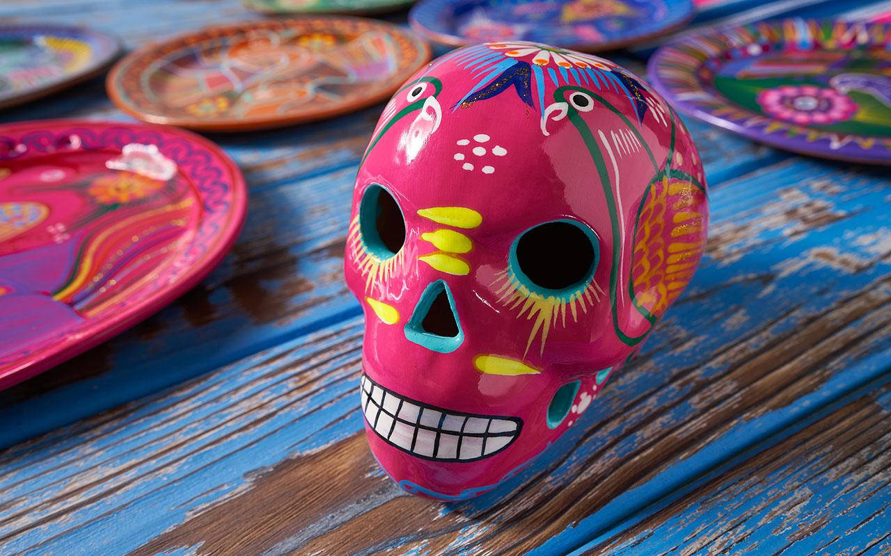Dia De Los Muertos 桌布 壁紙 — 鮮豔的粉紅色糖骷髏頭帶有彩色花卉圖案，放置在風化的藍色木桌上，周圍環繞著裝飾盤子。