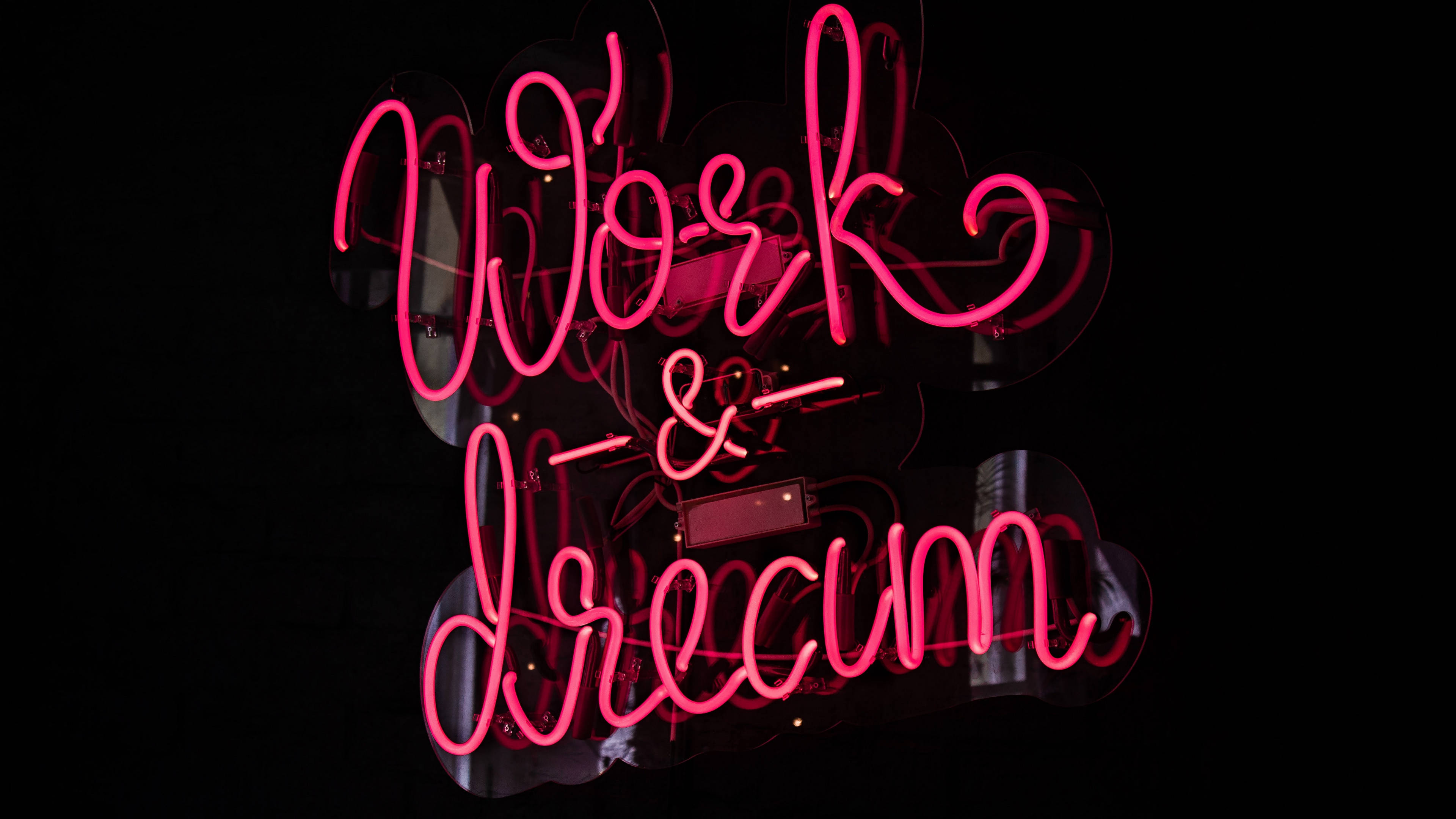 霓虹紅 桌布 壁紙 — 鮮豔的粉紅色霓虹燈牌，上面寫著「Work & Dream」，在深色反光表面上發光。