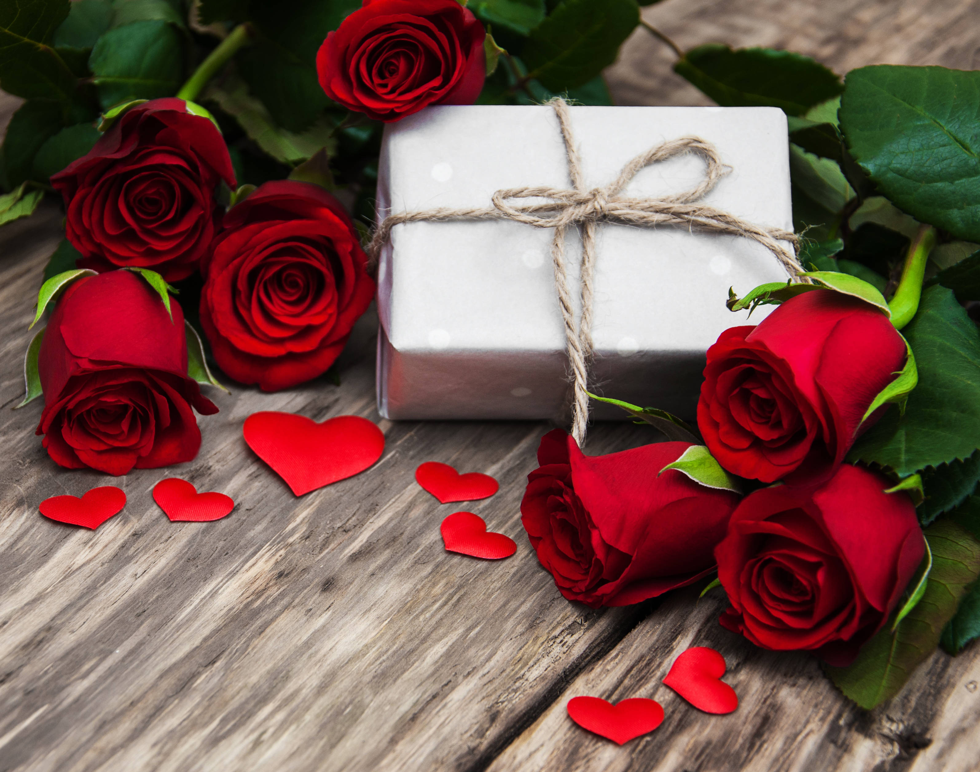 Romantic Rose 桌布 壁紙 — 鮮豔的紅玫瑰和紙心環繞著一個用麻繩綁著的銀色禮物盒，放在質樸的木質表面上。