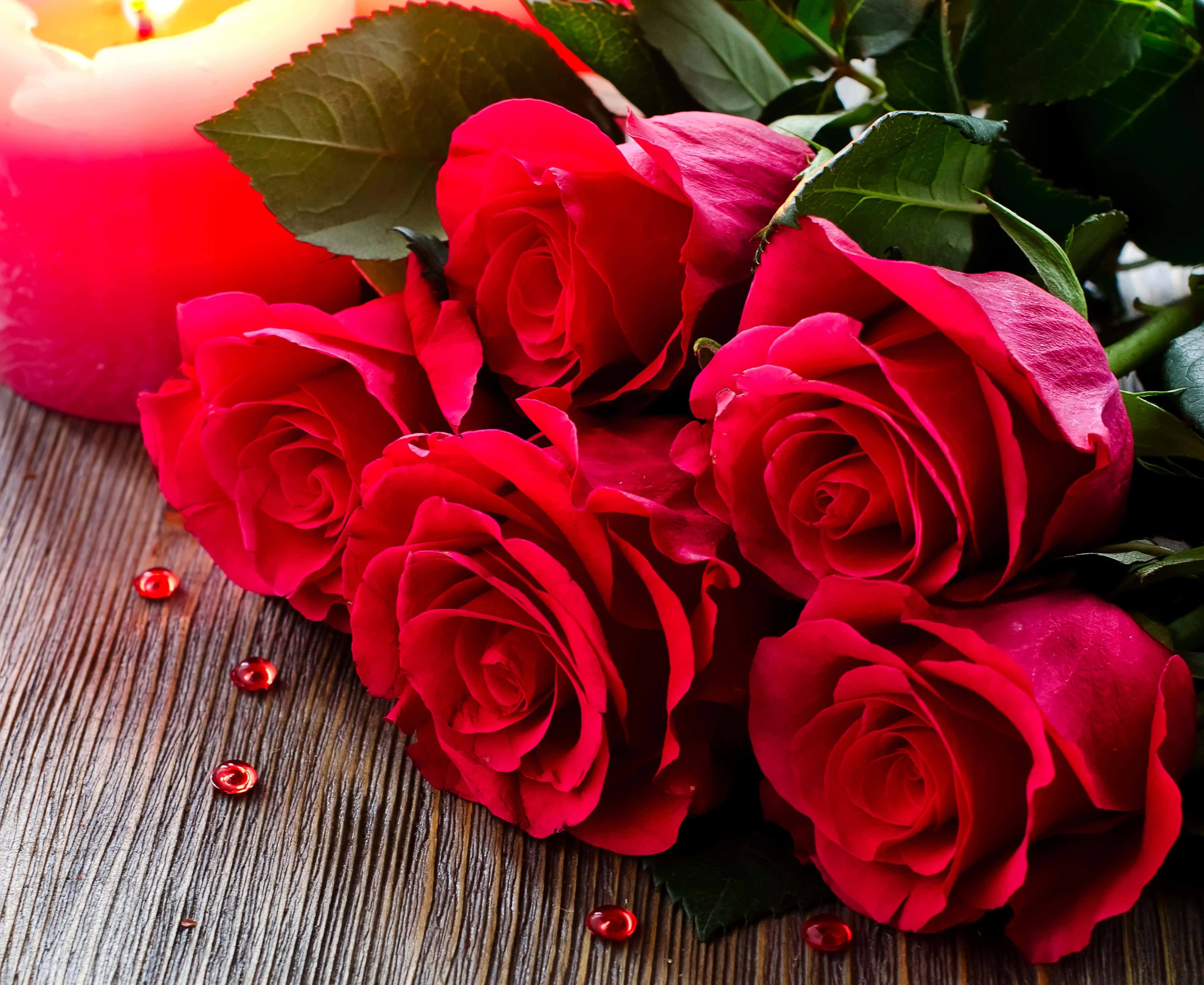 Bouquet 桌布 壁紙 — 鮮豔的紅玫瑰帶有露珠，旁邊放著一支發光的蠟燭，置於質樸的木質表面上。