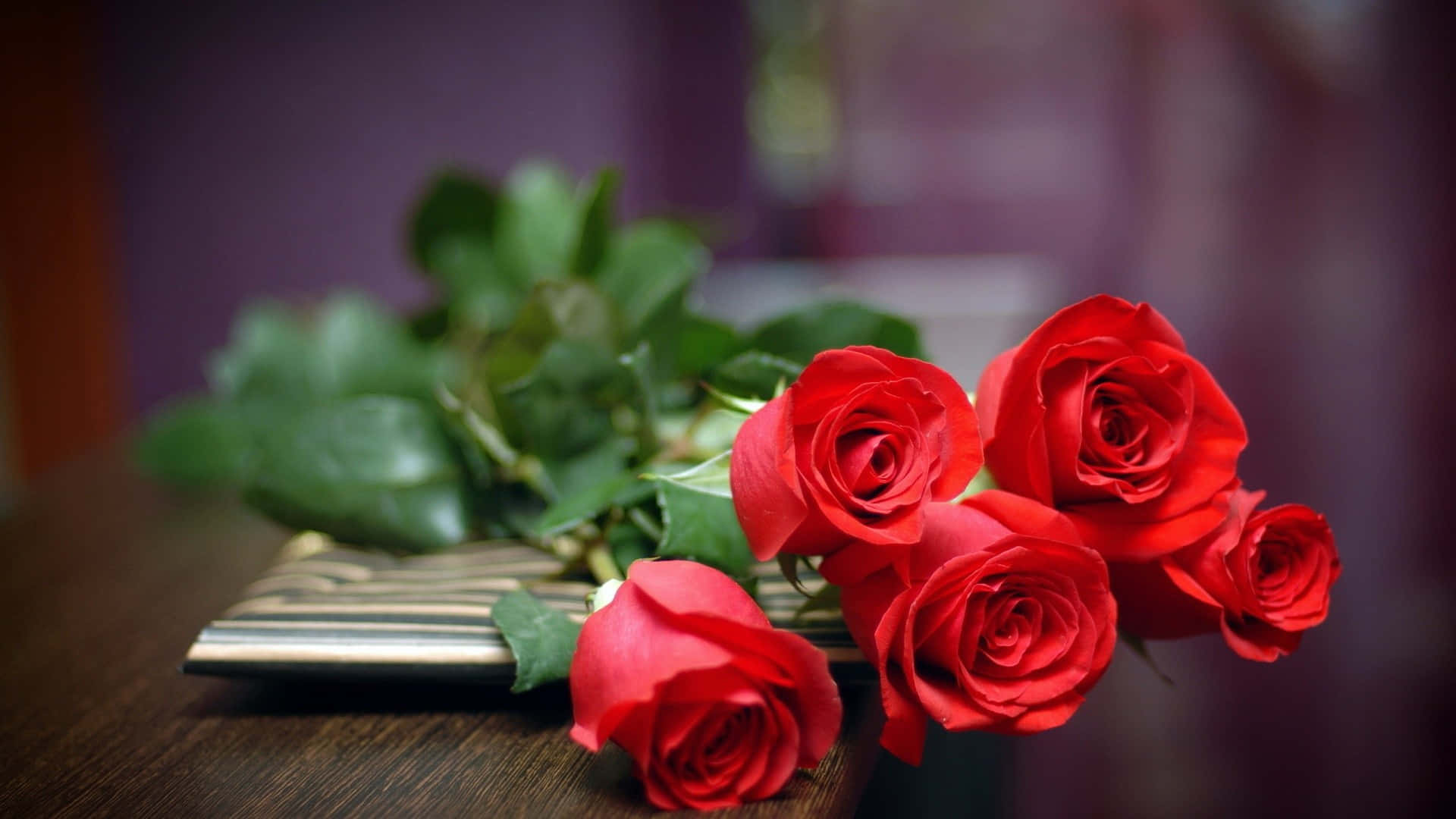 Cool Rose 桌布 壁紙 — 鮮豔的紅玫瑰與茂盛的綠葉優雅地放置在木桌上，旁邊有一本條紋筆記本。